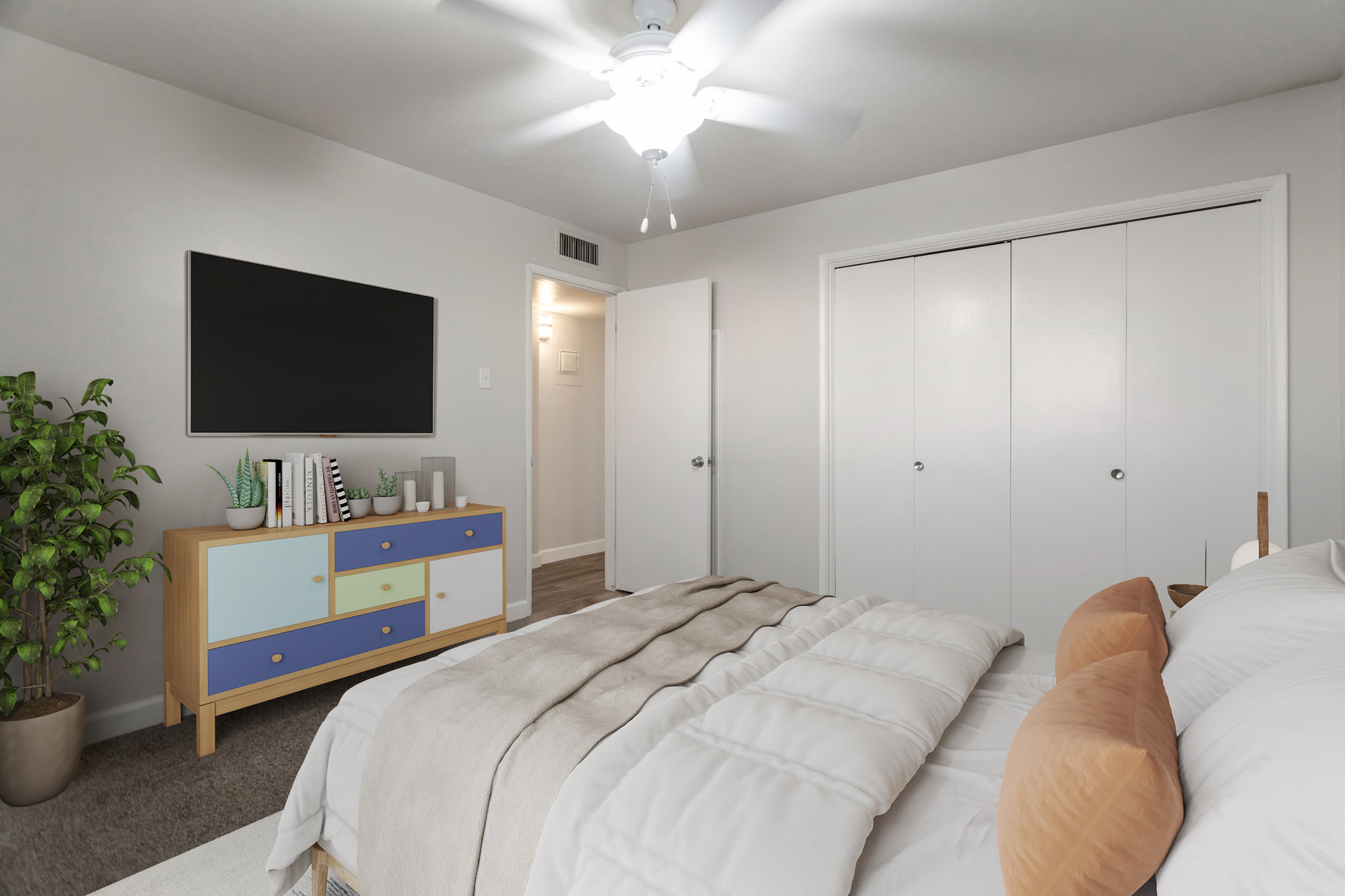 A modern bedroom featuring a bed with fluffy white bedding and two decorative pillows. There's a colorful dresser with geometric patterns, a flat-screen TV mounted on the wall, and a potted plant in the corner. Light from a ceiling fan illuminates the space, which has neutral-colored walls and a closet with sliding doors.