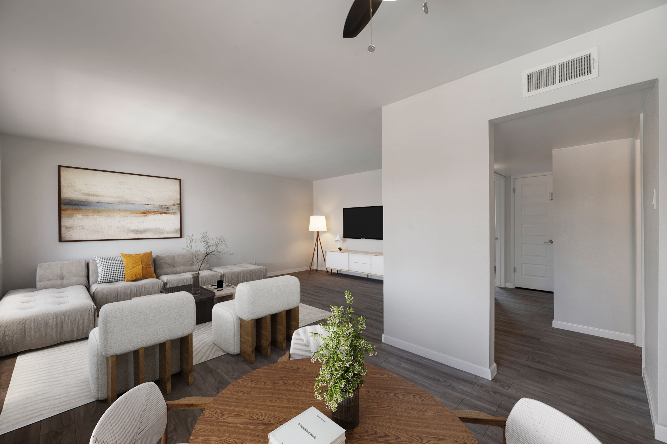 Modern living room featuring a light gray sectional sofa with a yellow throw pillow, a round wooden coffee table, and minimalist decor. A large wall-mounted TV is visible, along with a floor lamp providing soft lighting. A doorway leads to another room, while a plant adds a touch of greenery to the space.