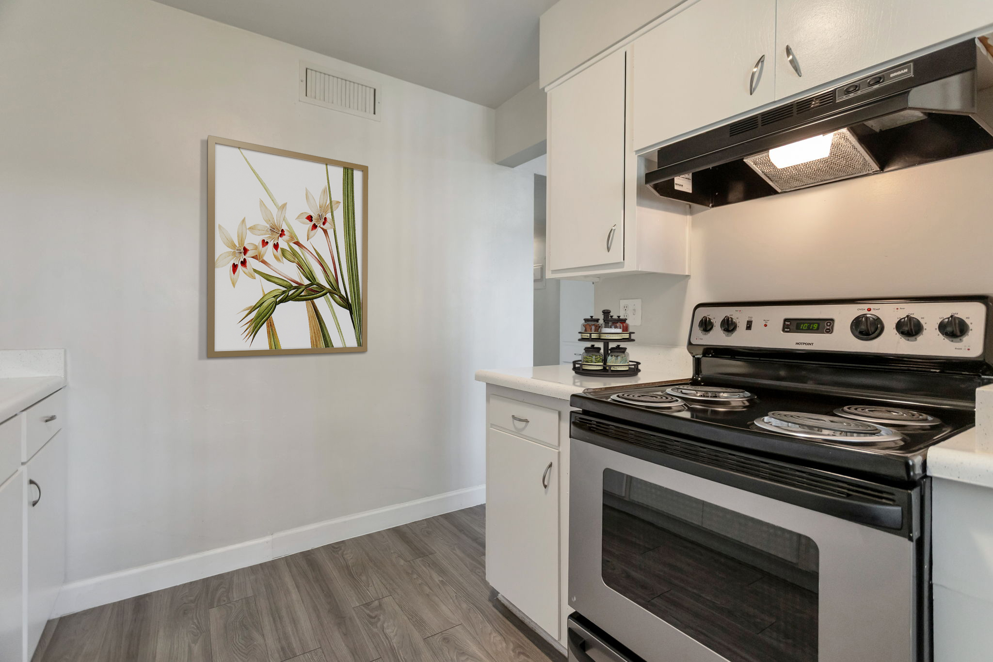A modern kitchen featuring a black stove, white cabinets, and a sleek countertop. A colorful artwork of orchids is hung on the wall, adding a touch of elegance. The floor is made of gray wood, and the overall ambiance is bright and inviting.