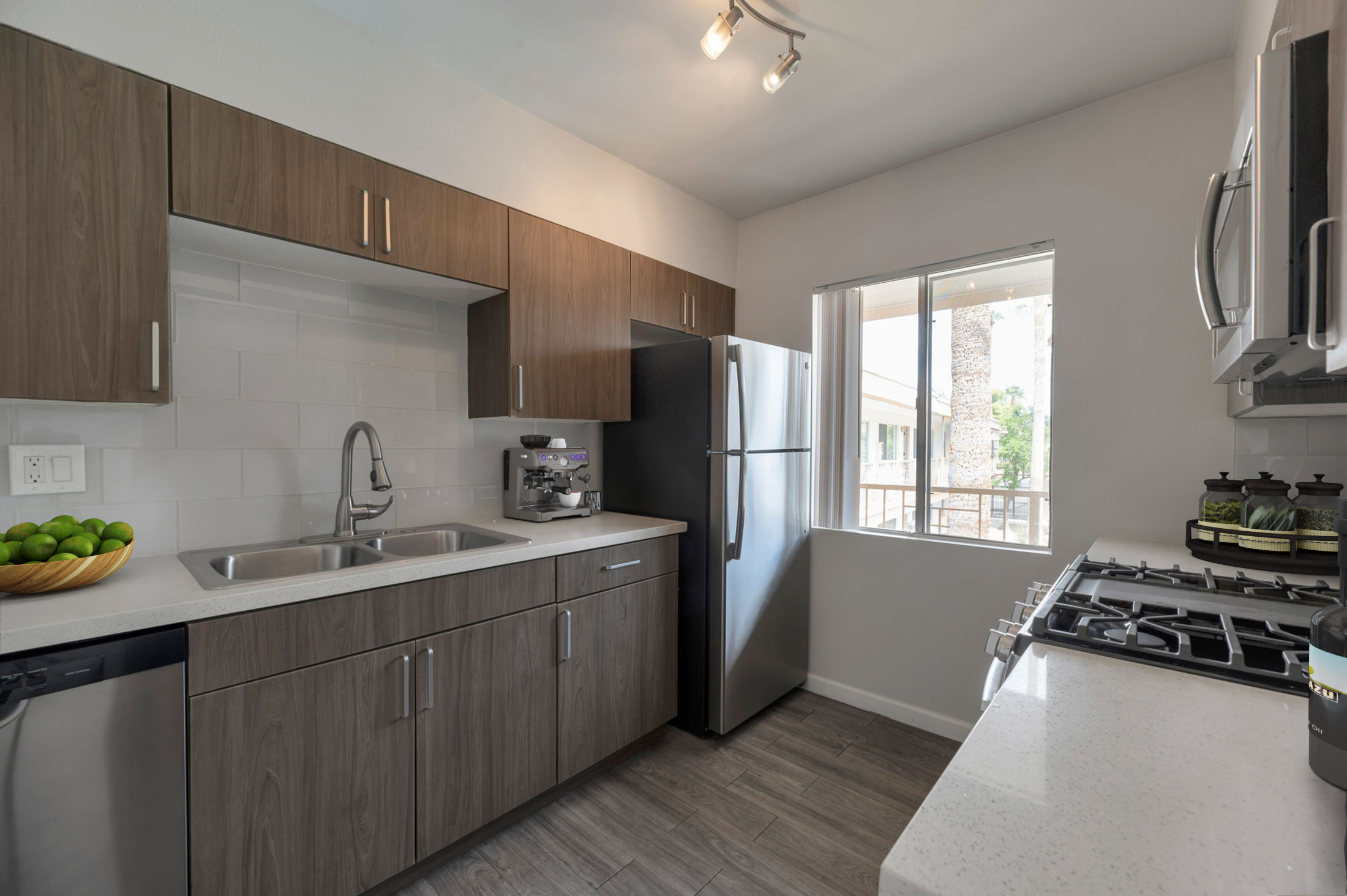 Modern kitchen featuring wooden cabinets, a stainless steel refrigerator, and a double sink. Countertop made of light-colored stone. A bowl of limes sits on the counter, and a window allows natural light to brighten the space. Stainless steel appliances complement the sleek design.