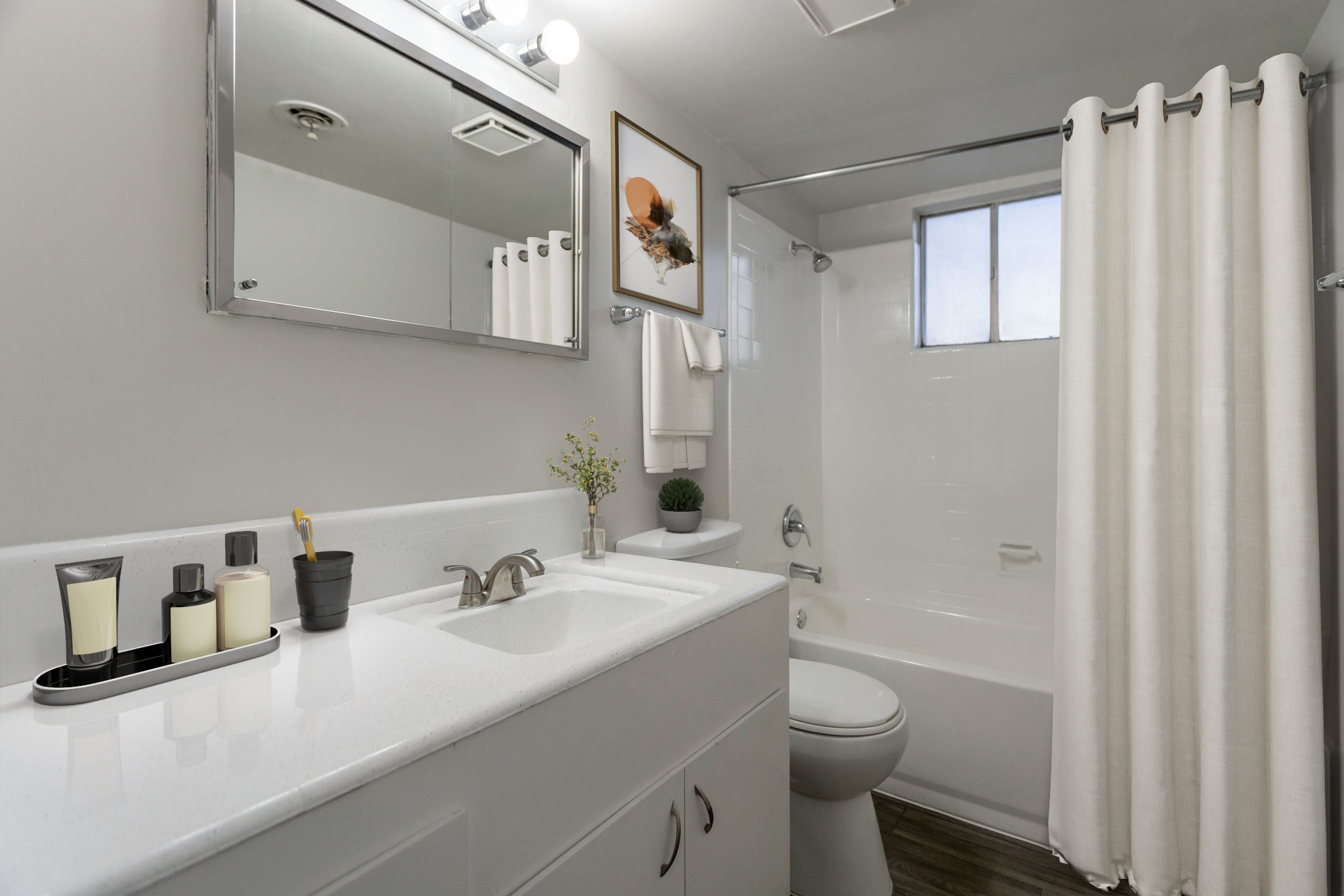 A modern bathroom featuring a white countertop with toiletries, a clean bathtub with a white shower curtain, a mirror above the sink, and a window allowing natural light. Decorative elements include a small plant and a framed picture on the wall.