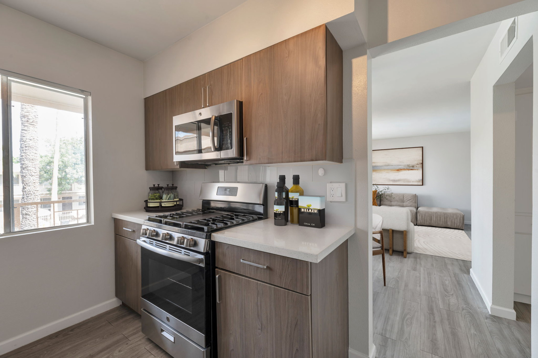 Modern kitchen featuring wooden cabinets, a sleek stove and oven, stainless steel microwave, and countertop. A large window allows natural light, revealing a glimpse of the outdoors. In the background, a living area with a gray couch is visible.