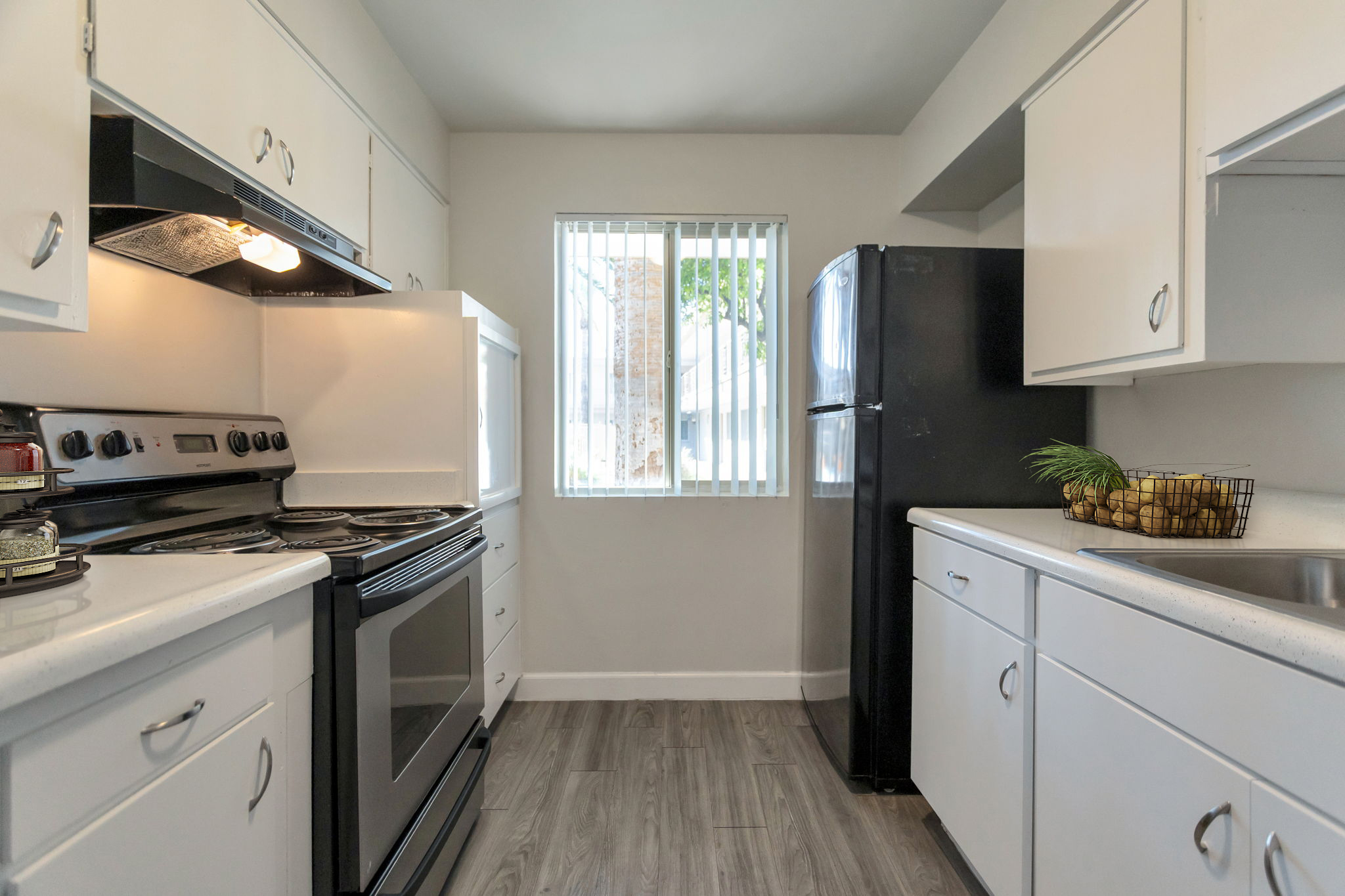 A bright, modern kitchen featuring white cabinets, a black refrigerator, and an electric stove with an overhead vent. A window with vertical blinds offers natural light, and the floor has a wood-like finish. A basket of fruit adds a decorative touch on the countertop.