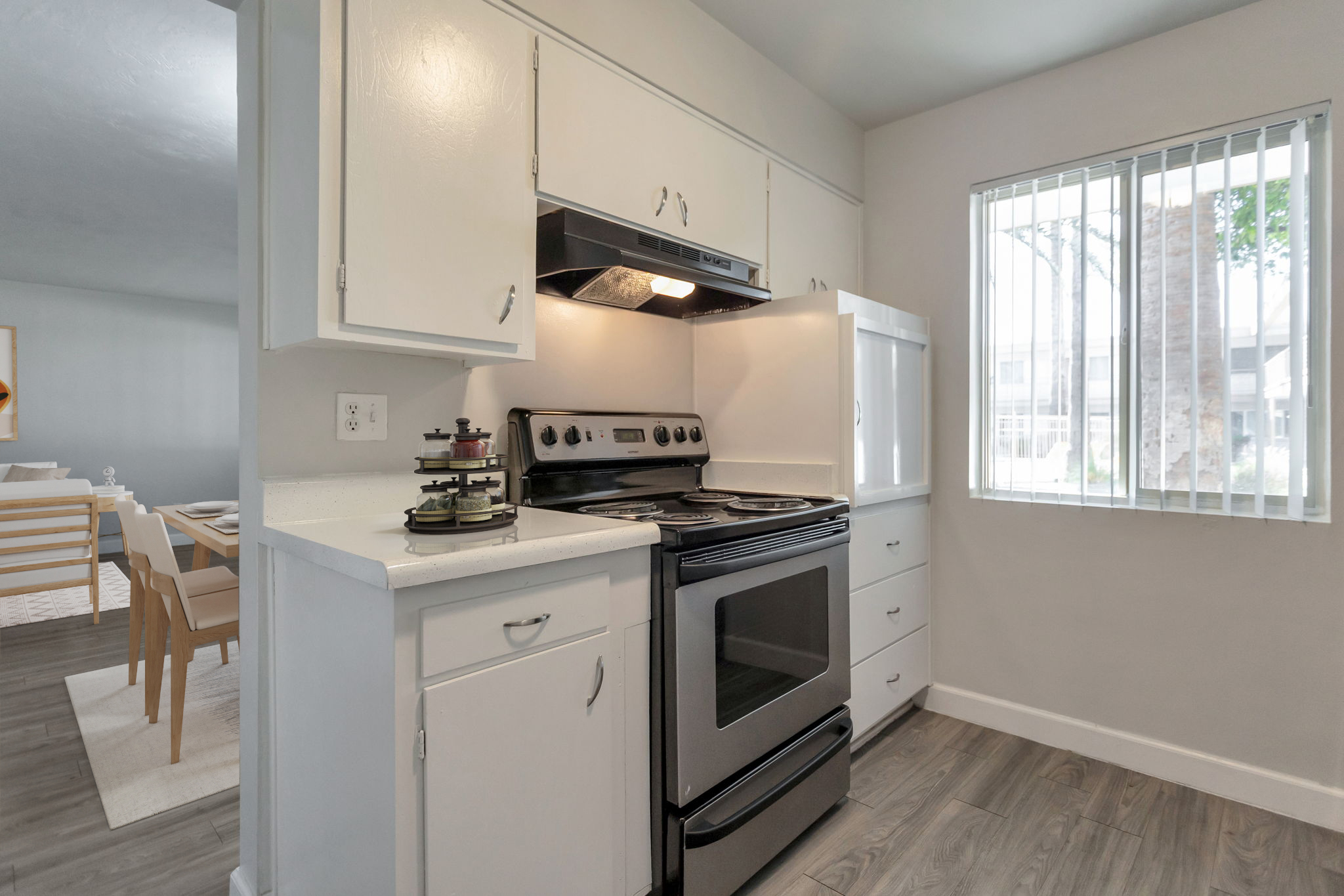 A modern kitchen featuring a black stovetop and oven, white cabinetry, and a countertop. There is a small shelf with spices or condiments beside the stove, and a window with horizontal blinds allows natural light to enter the space. The flooring is a light wood tone, creating a clean and inviting atmosphere.