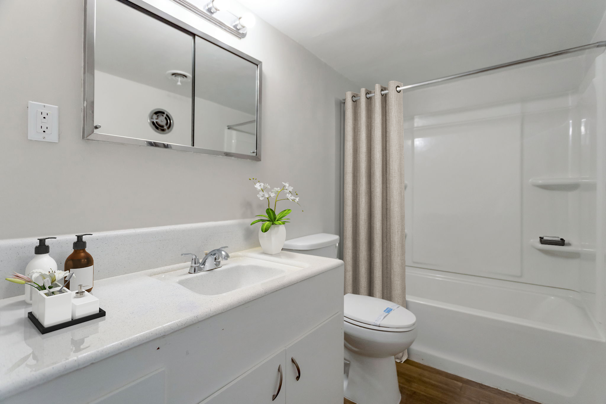 A clean, modern bathroom featuring a white vanity with a sink, two decorative bottles, and a small potted plant. The shower area has a light-colored curtain and the walls are painted a neutral tone. A mirror is mounted above the sink, and the flooring is wooden.