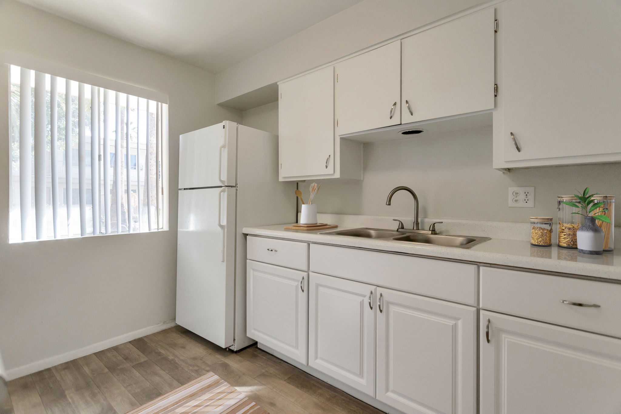 Bright kitchen featuring white cabinets and a light countertop. A stainless steel sink is between two burners, with utensils and a plant on the counter. A refrigerator is on the left, and a window with vertical blinds allows natural light to illuminate the space.