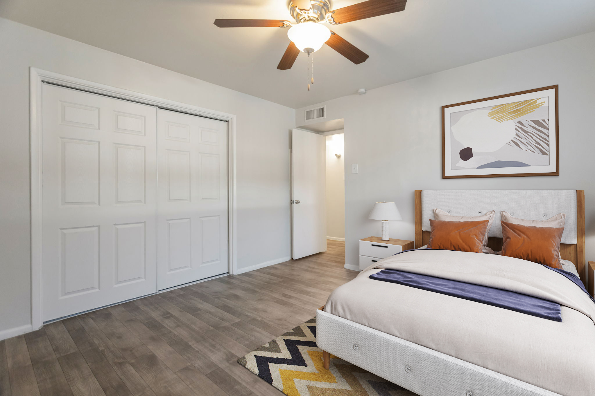 A well-lit bedroom featuring a bed with decorative pillows, a modern ceiling fan, and a light-colored rug. There's a nightstand with a lamp next to the bed, and a large closet with double doors. A minimalist piece of art hangs on the wall, enhancing the room's contemporary design.