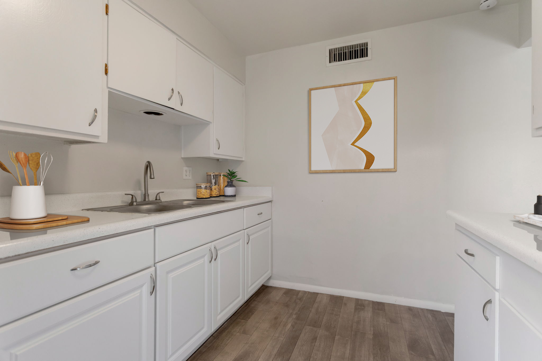 A modern kitchen featuring white cabinets, a stainless steel sink, and a light-colored countertop. The walls are painted in a neutral tone, and a piece of abstract wall art with gold accents hangs above the sink. A small plant and kitchen utensils are placed on the counter, adding a touch of decor.