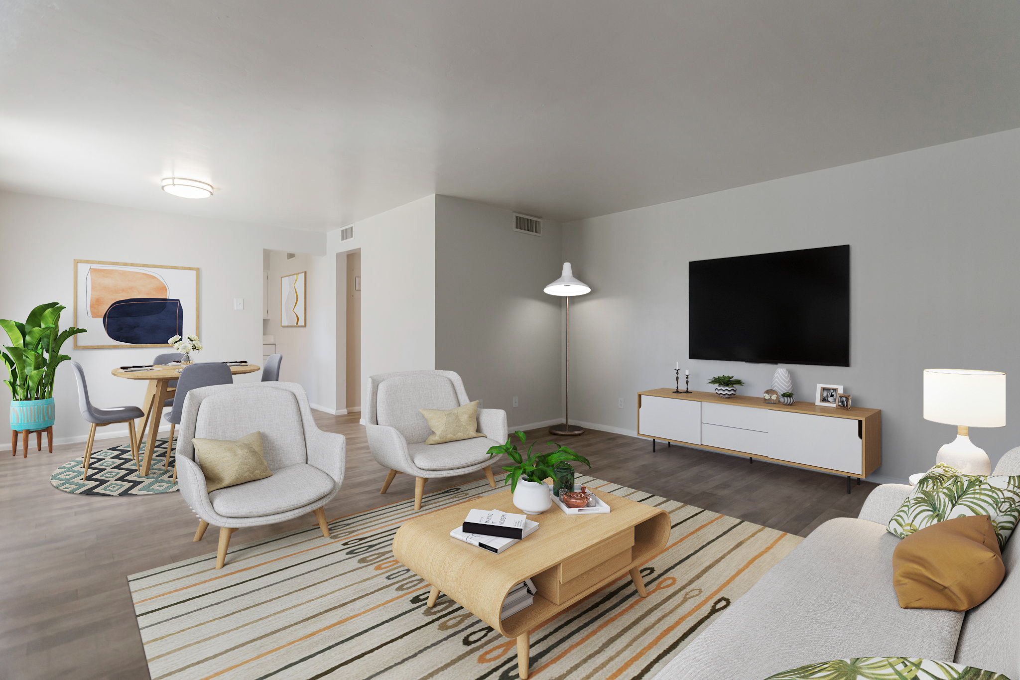 A modern living room featuring two light gray armchairs and a beige coffee table on a striped area rug. A white sofa with decorative pillows is against a wall, alongside a TV unit with a flat-screen TV. A dining area is visible in the background with a round table and chairs, and a large indoor plant adds greenery to the space.