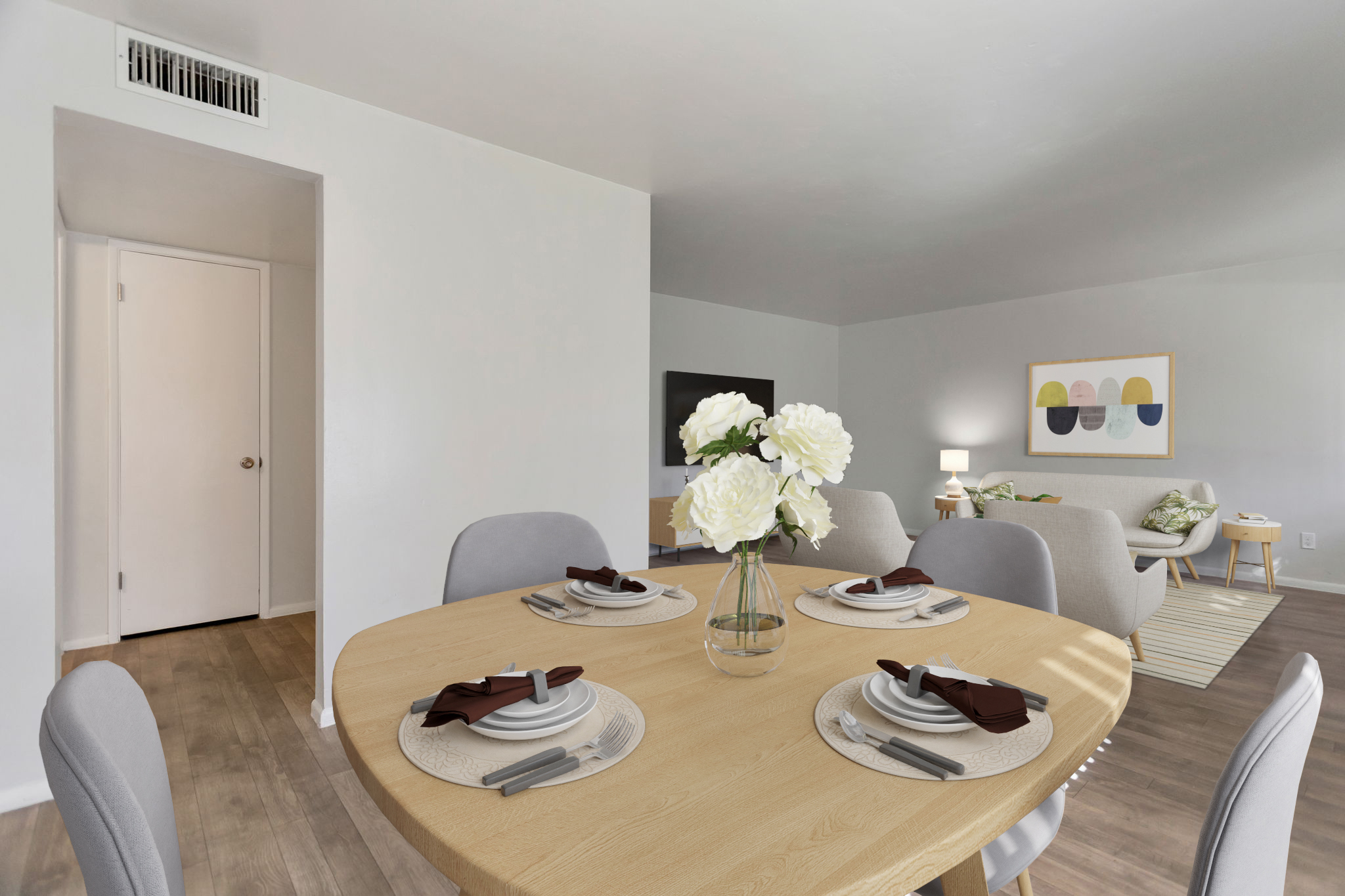 A modern dining area featuring a round wooden table set for four with plates, utensils, and a floral centerpiece. The room has light-colored walls, hardwood flooring, and nearby furniture indicating a cozy living space. A door leads to another room in the background, with soft lighting enhancing the ambiance.