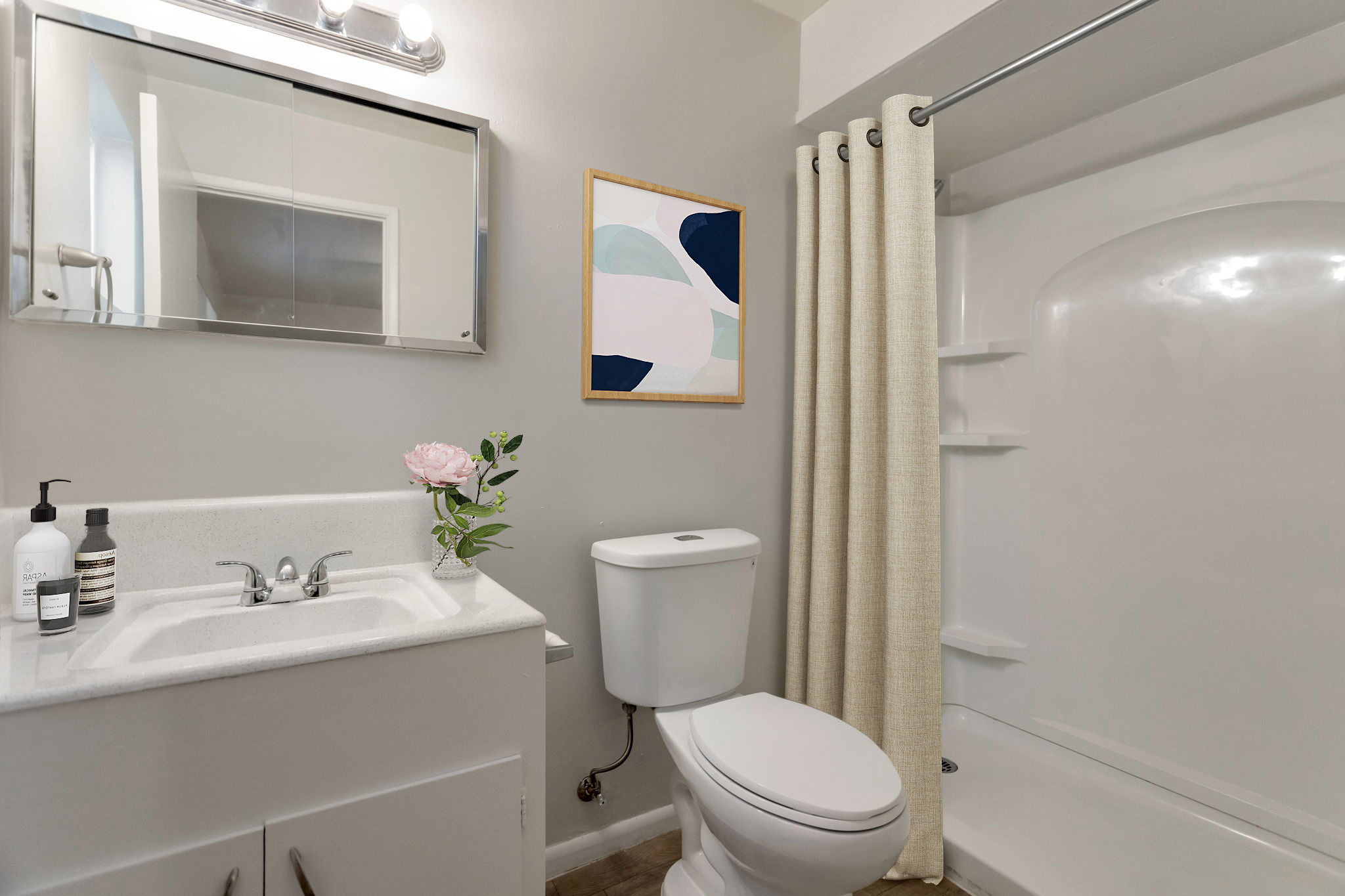 A modern bathroom featuring a white sink with a mirror above, a toilet, and a shower area with a clear curtain. There's a vase with a pink flower on the sink and abstract artwork on the wall, complemented by neutral-colored walls and flooring. The overall decor is clean and contemporary.