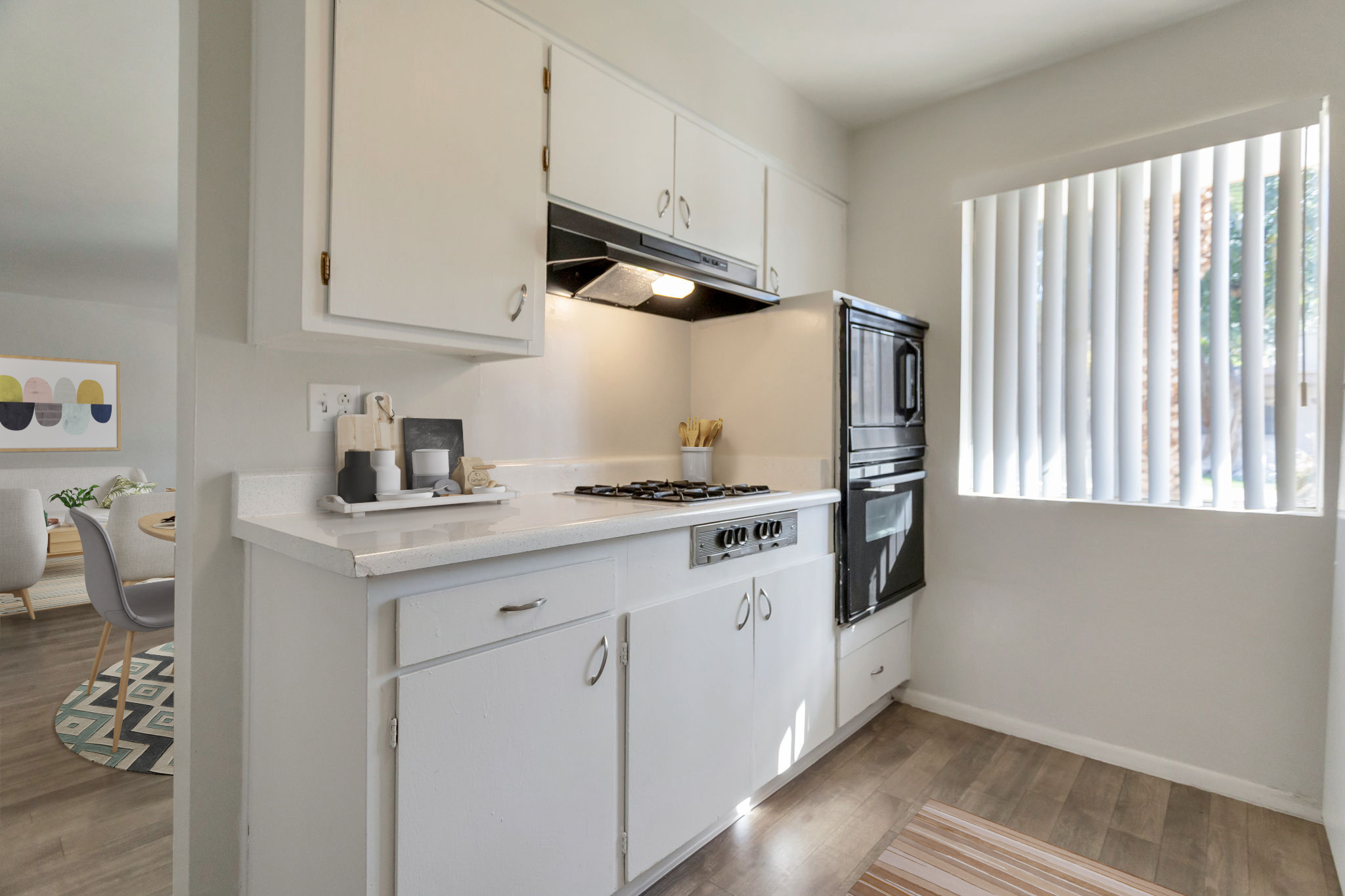 A modern kitchen with white cabinets and a black oven. The countertop features dishware and utensils. A large window with vertical blinds allows natural light to enter, illuminating the space. Light wooden flooring and a striped rug add warmth to the room. A dining area is visible in the background.