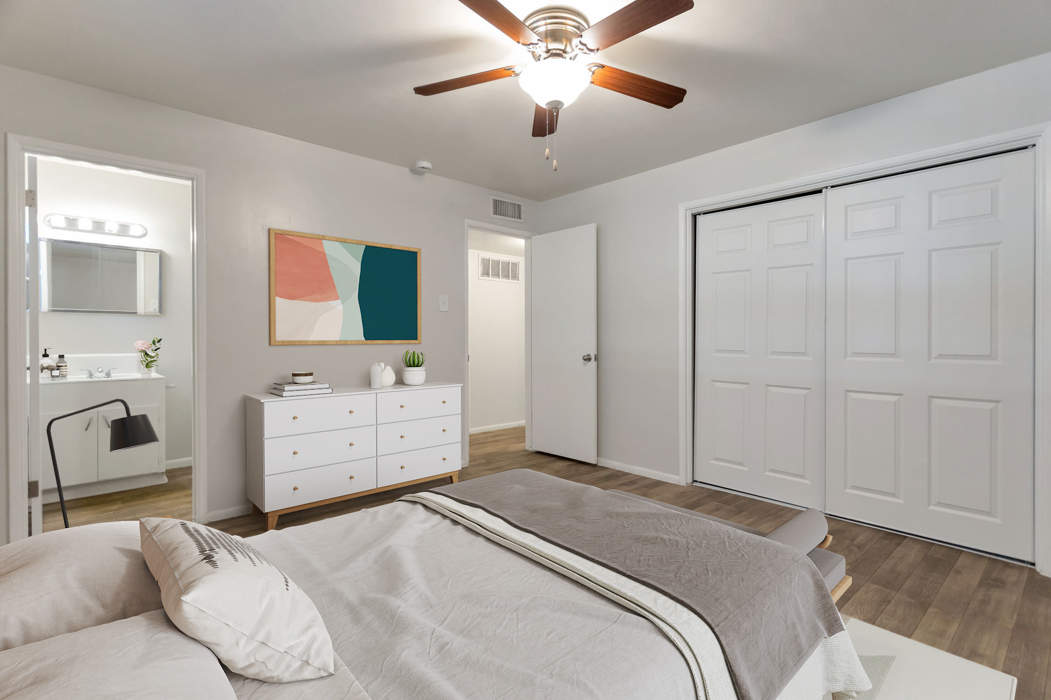 A modern bedroom featuring a bed with a light gray blanket, a white dresser, and a ceiling fan. In the background, a bathroom door is visible along with a painting on the wall. There are two closet doors, and the room has wood-like flooring and neutral-colored walls.