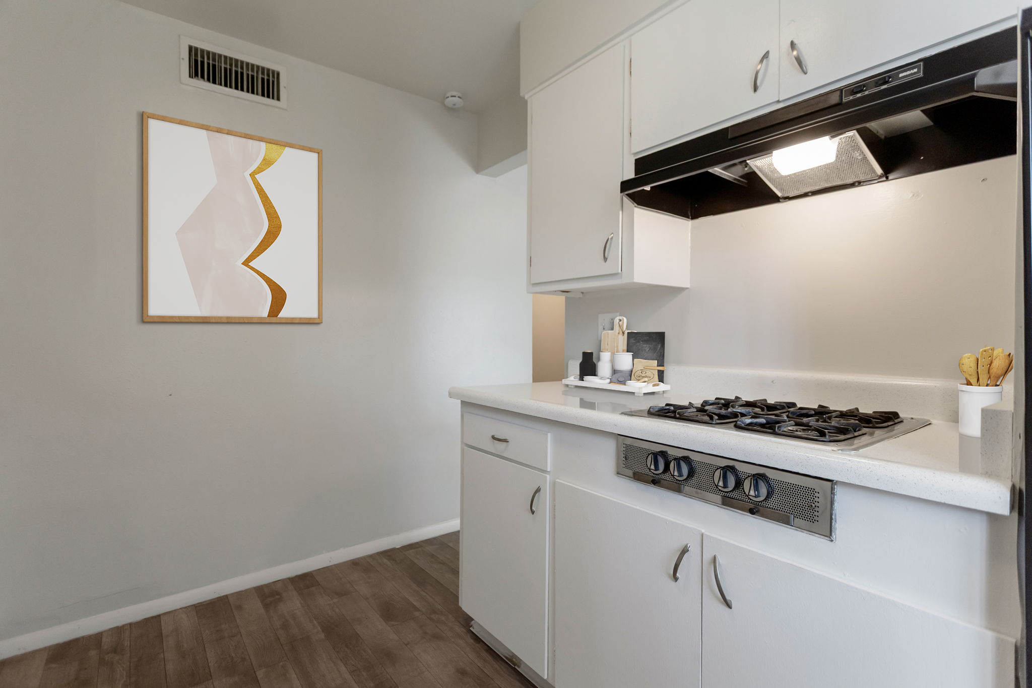 Modern kitchen featuring a white countertop, a gas stove with a black hood, and minimalist cabinetry. A framed abstract artwork with a gold accent is hanging on the wall. The floor is a warm wood-tone, creating a bright and inviting atmosphere.
