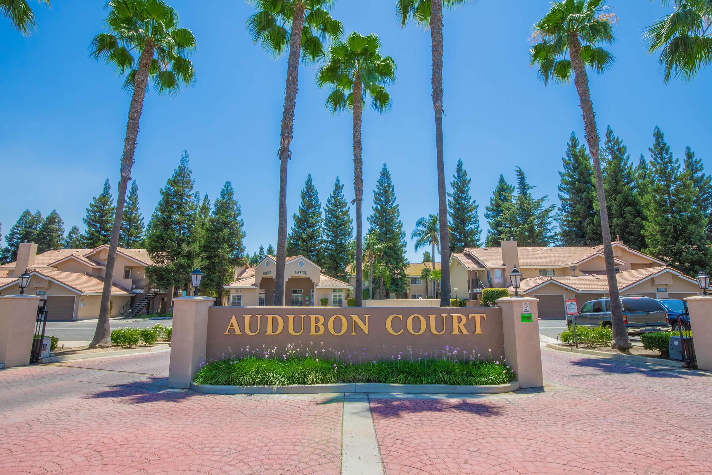 Entrance to Audubon Court, featuring a large sign with "Audubon Court" in bold letters. The area is surrounded by tall palm trees and lush greenery, set against a clear blue sky. The driveway is paved with red bricks, leading into a residential complex with buildings in the background.