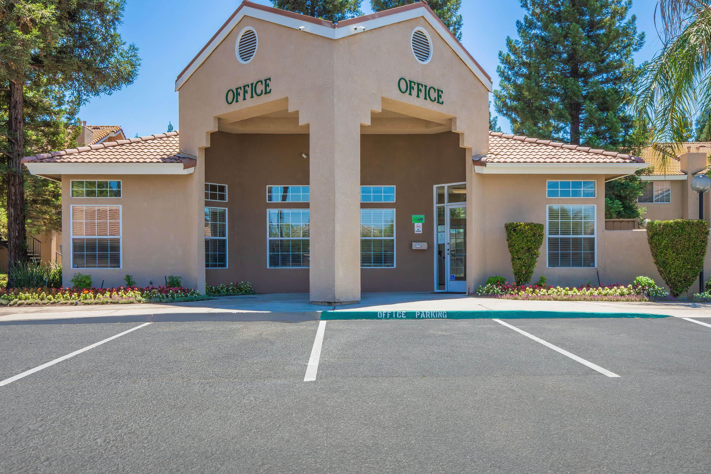 Two-story building with a Mediterranean architectural style. The building has a peaked roof, light brown exterior, and large windows. The word "OFFICE" is prominently displayed above the entrance. Surrounding landscaping includes green shrubs and flowers. The parking lot is visible in front of the building.