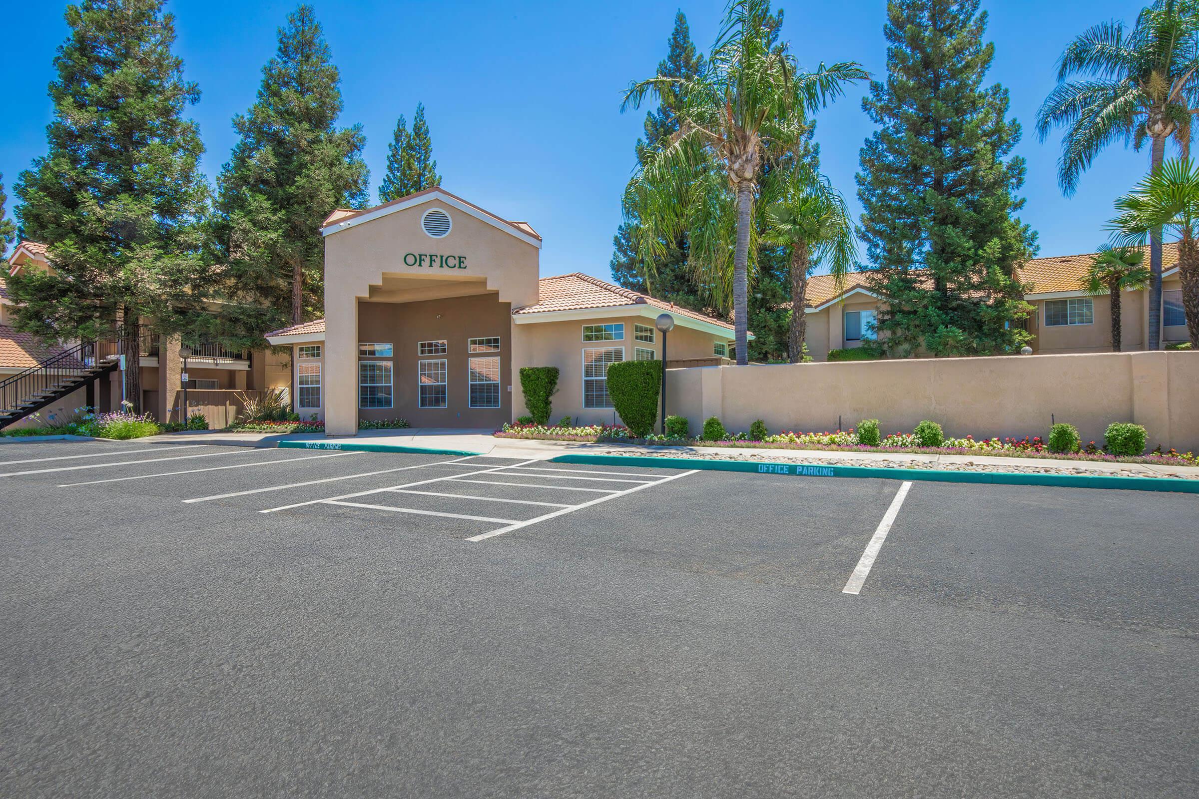 A well-maintained office building surrounded by palm trees and shrubs, situated in a parking lot. The building features a sign labeled "OFFICE" above the entrance. The clear blue sky and sunlight create a bright and welcoming atmosphere.