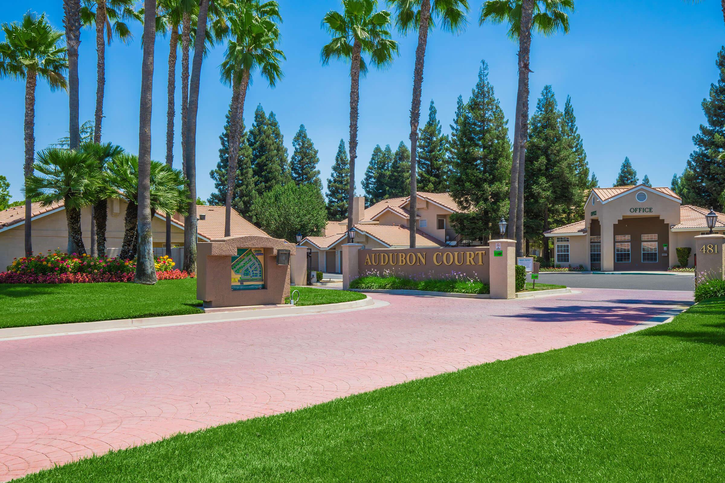 View of Audubon Court entrance with a welcoming sign, surrounded by well-maintained landscaping, including palm trees and colorful flower beds. There is a clear blue sky overhead and residential buildings visible in the background, creating a peaceful and inviting atmosphere.