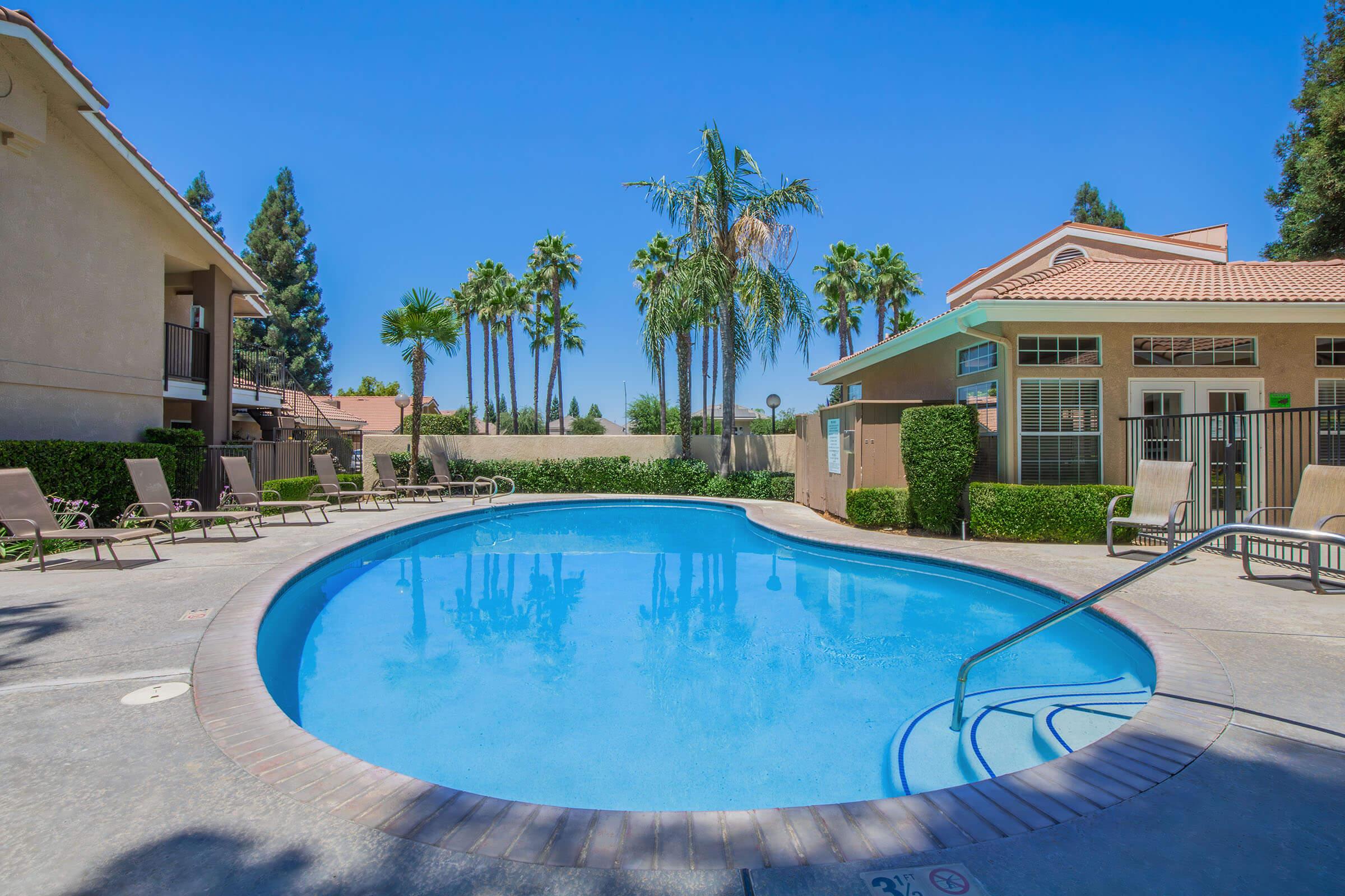 A serene swimming pool surrounded by palm trees and lounge chairs, with a light blue sky overhead. The pool is set within a residential area, featuring well-maintained landscaping and nearby buildings. The scene conveys a relaxing and inviting atmosphere for leisure and enjoyment.
