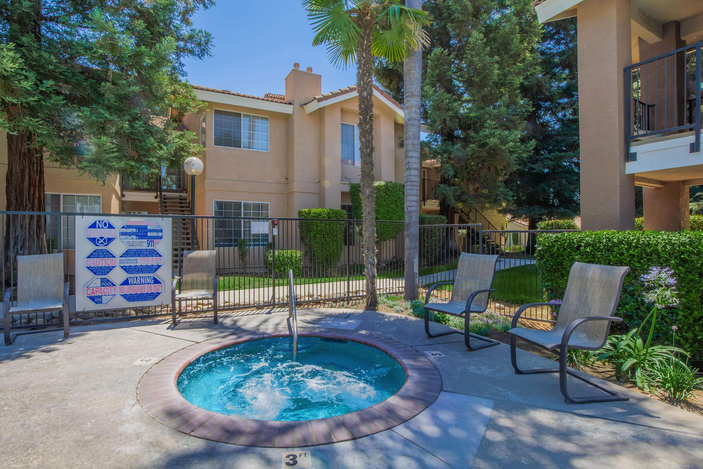 A hot tub surrounded by a fenced area, with two chairs nearby. Lush greenery and palm trees provide shade, and apartment buildings are visible in the background on a sunny day. A sign indicating pool rules is attached to the fence.