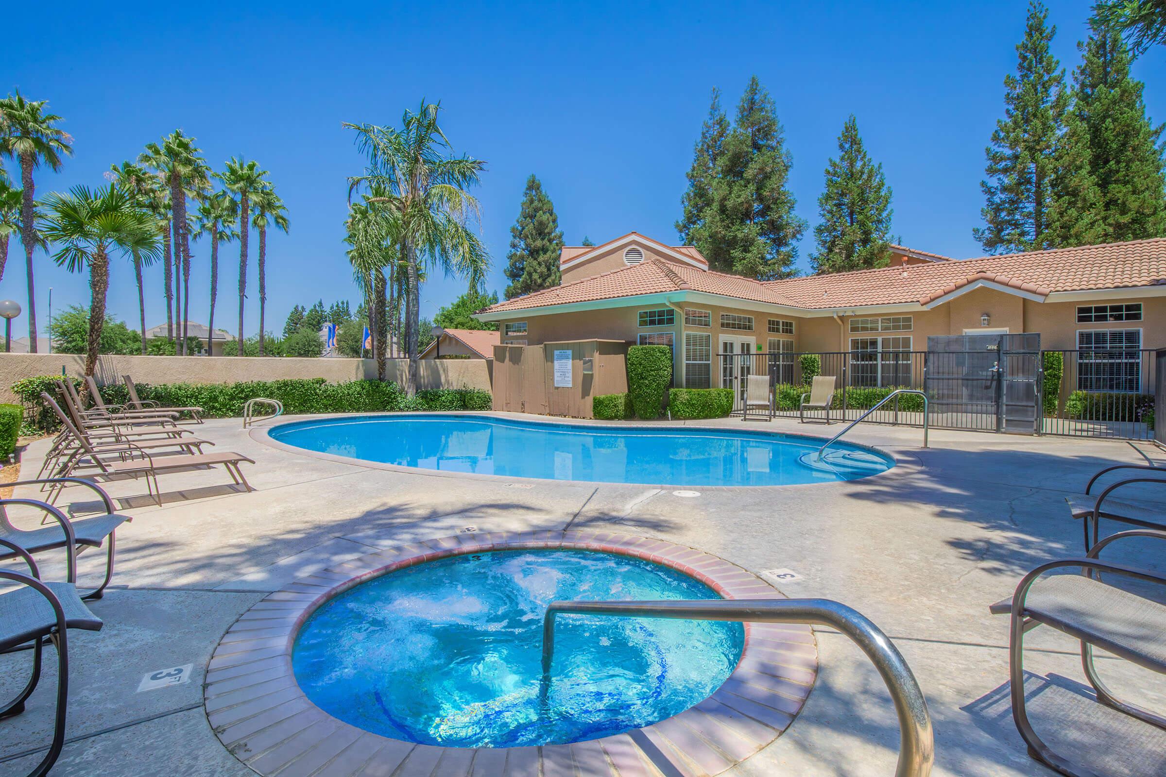 A sunny outdoor pool area featuring a large swimming pool and a hot tub, surrounded by palm trees and greenery. There are lounge chairs arranged around the pool, and a clubhouse with windows in the background. The clear blue sky enhances the relaxing atmosphere of the space.