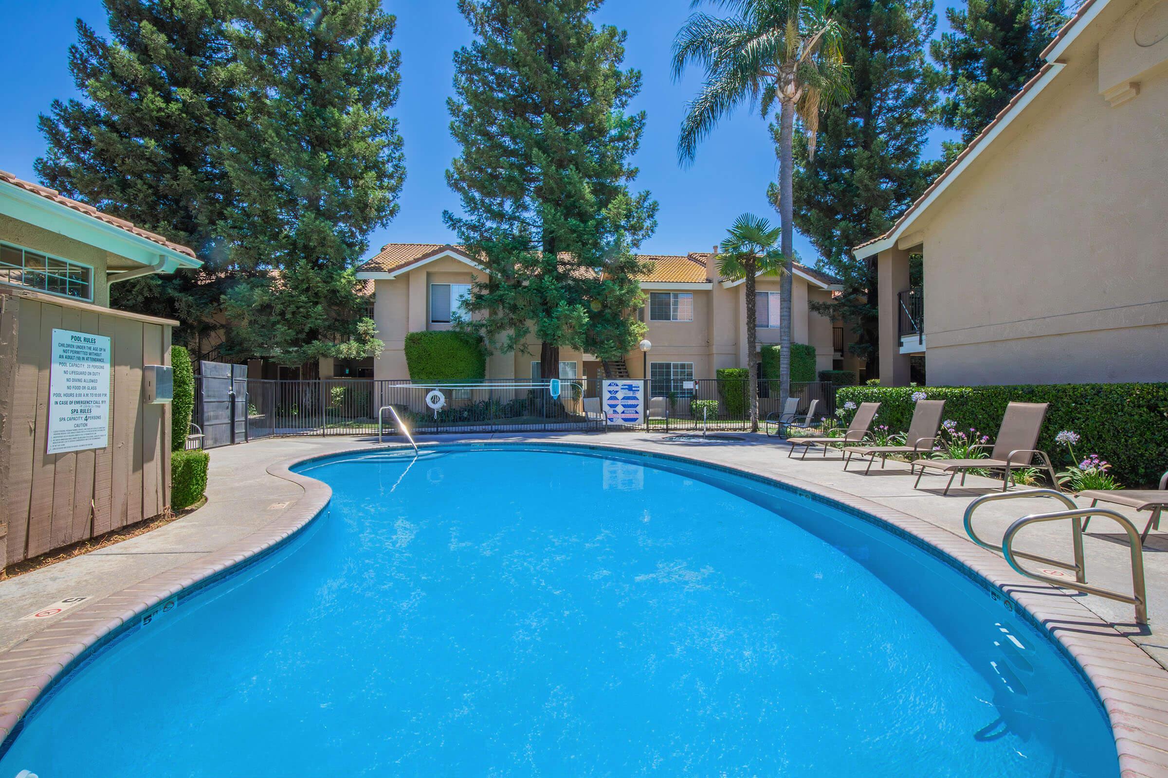 A clear blue swimming pool surrounded by lush green trees and well-maintained landscaping. Comfortable lounge chairs are positioned by the poolside, and nearby, a privacy fence encloses the area. Bright sunlight creates a cheerful atmosphere, highlighting the residential buildings in the background.