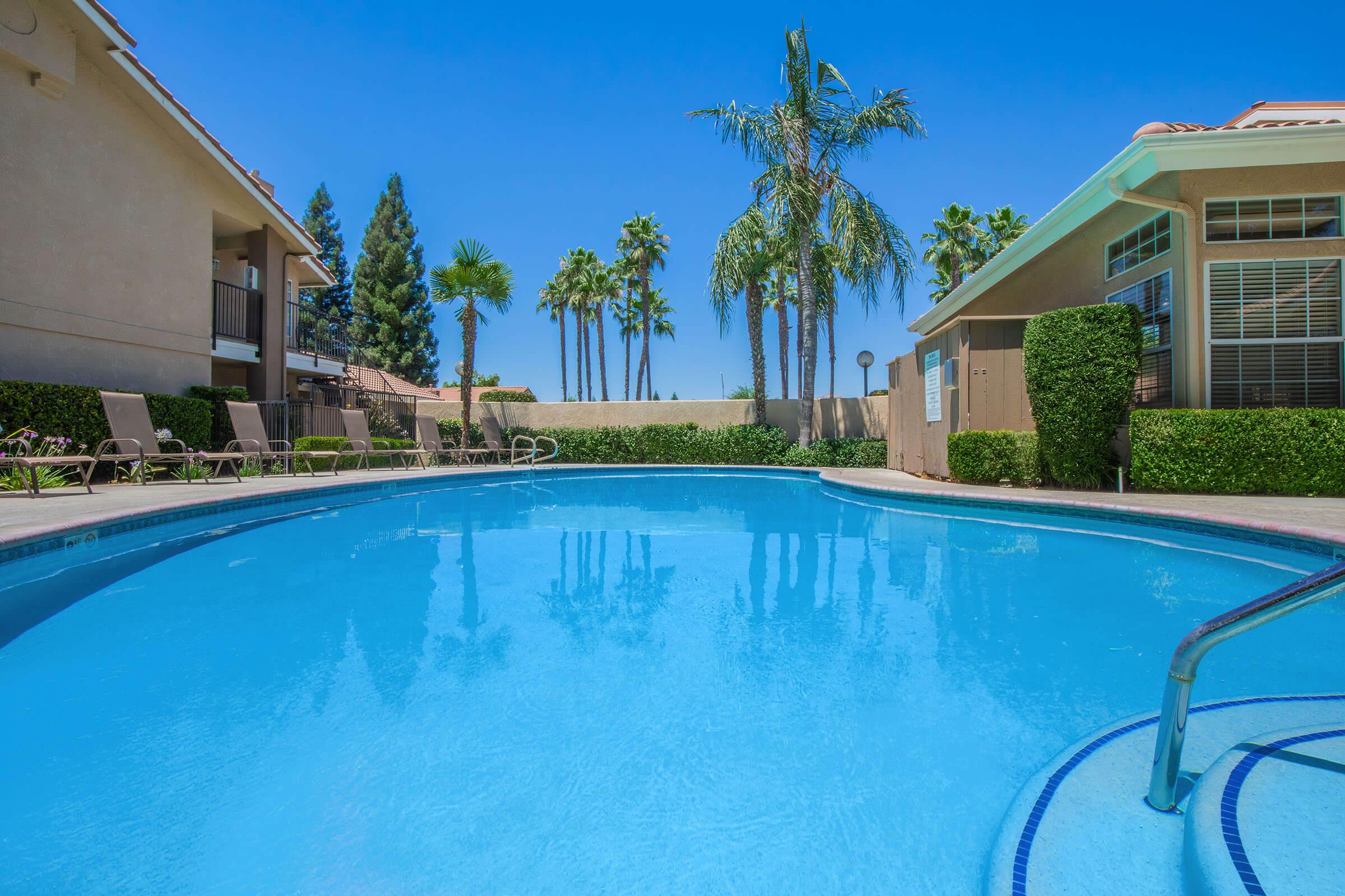 A clear blue swimming pool surrounded by palm trees and lounge chairs. The sun shines brightly in a clear blue sky, creating a relaxing atmosphere. Nearby buildings feature balconies, adding to the inviting setting perfect for leisure and relaxation.