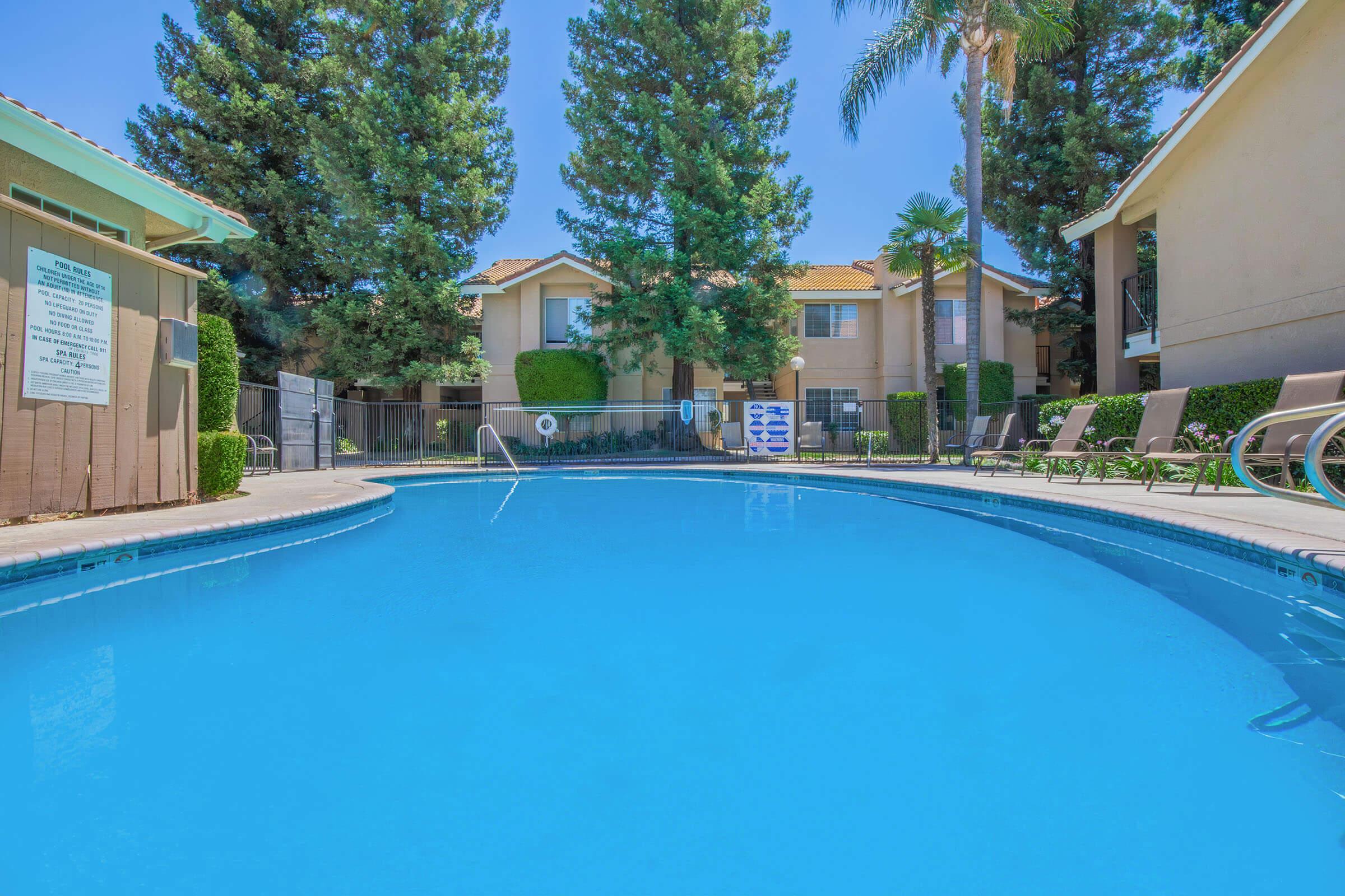 A clear blue swimming pool in an outdoor area surrounded by tall trees and apartment buildings. Lounge chairs are positioned around the pool, and a sign is visible near the pool area, indicating safety rules or regulations. Bright sunshine creates a vibrant, inviting atmosphere.
