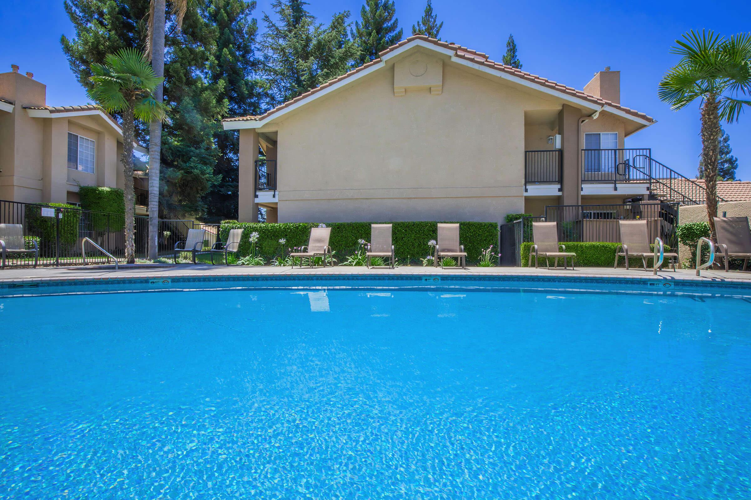A clear blue swimming pool reflecting a bright sky, surrounded by lounge chairs. In the background, a two-story building with a balcony is nestled among tall trees and well-maintained hedges, creating a peaceful and inviting atmosphere.