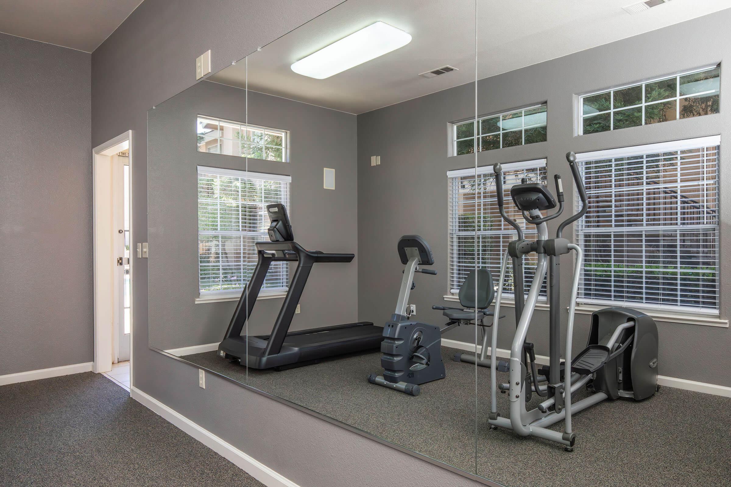 A modern gym interior featuring a treadmill, an elliptical machine, and a stationary bike. Large windows with white blinds let in natural light, and a full-length mirror reflects the workout equipment. The walls are painted a neutral gray, creating a calm atmosphere for exercise.
