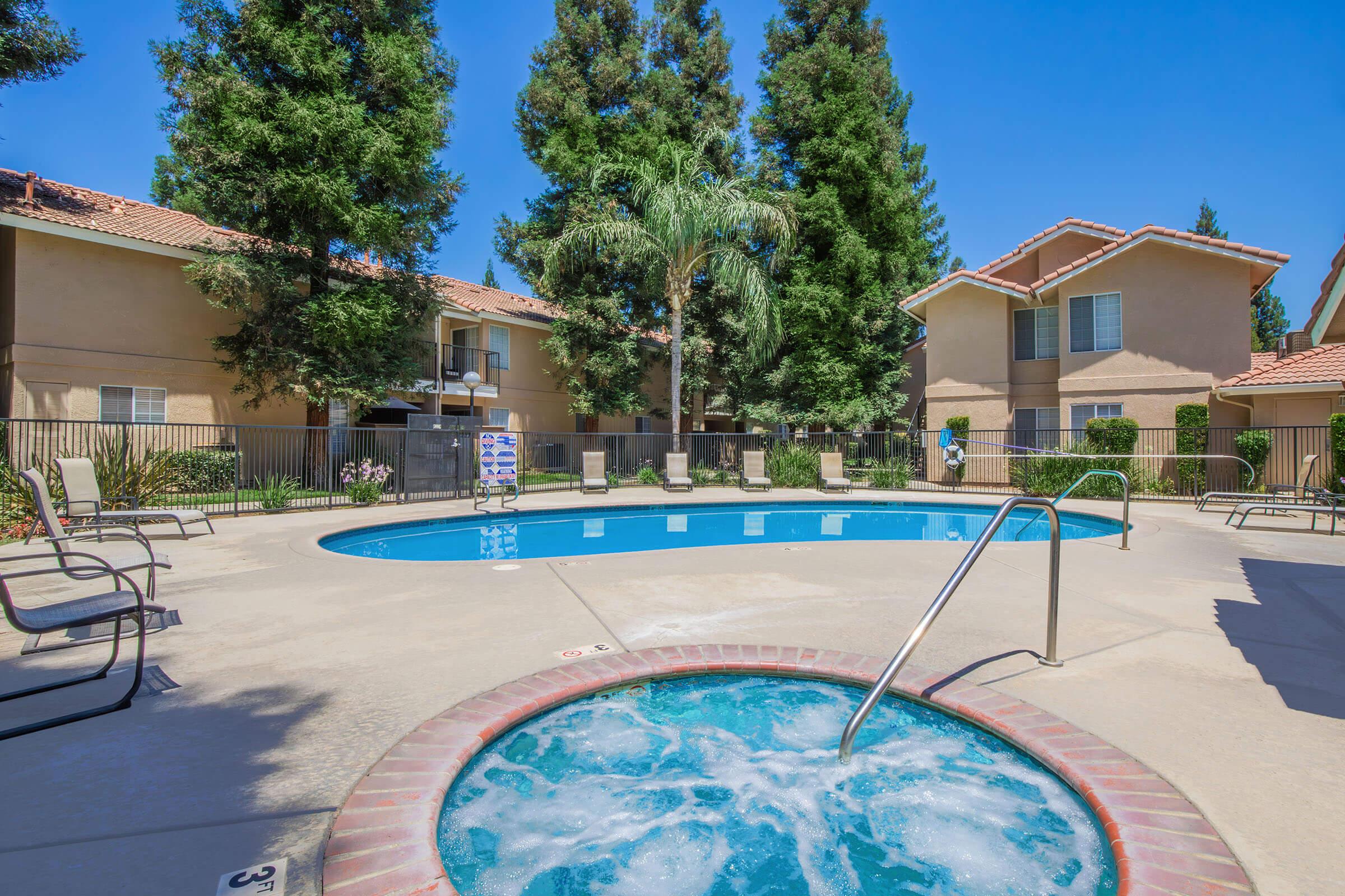 A sunny outdoor pool area featuring a blue swimming pool surrounded by lounge chairs and palm trees. A hot tub is situated nearby, with apartment buildings in the background. The space is well-maintained and invites relaxation in a bright, cheerful atmosphere.