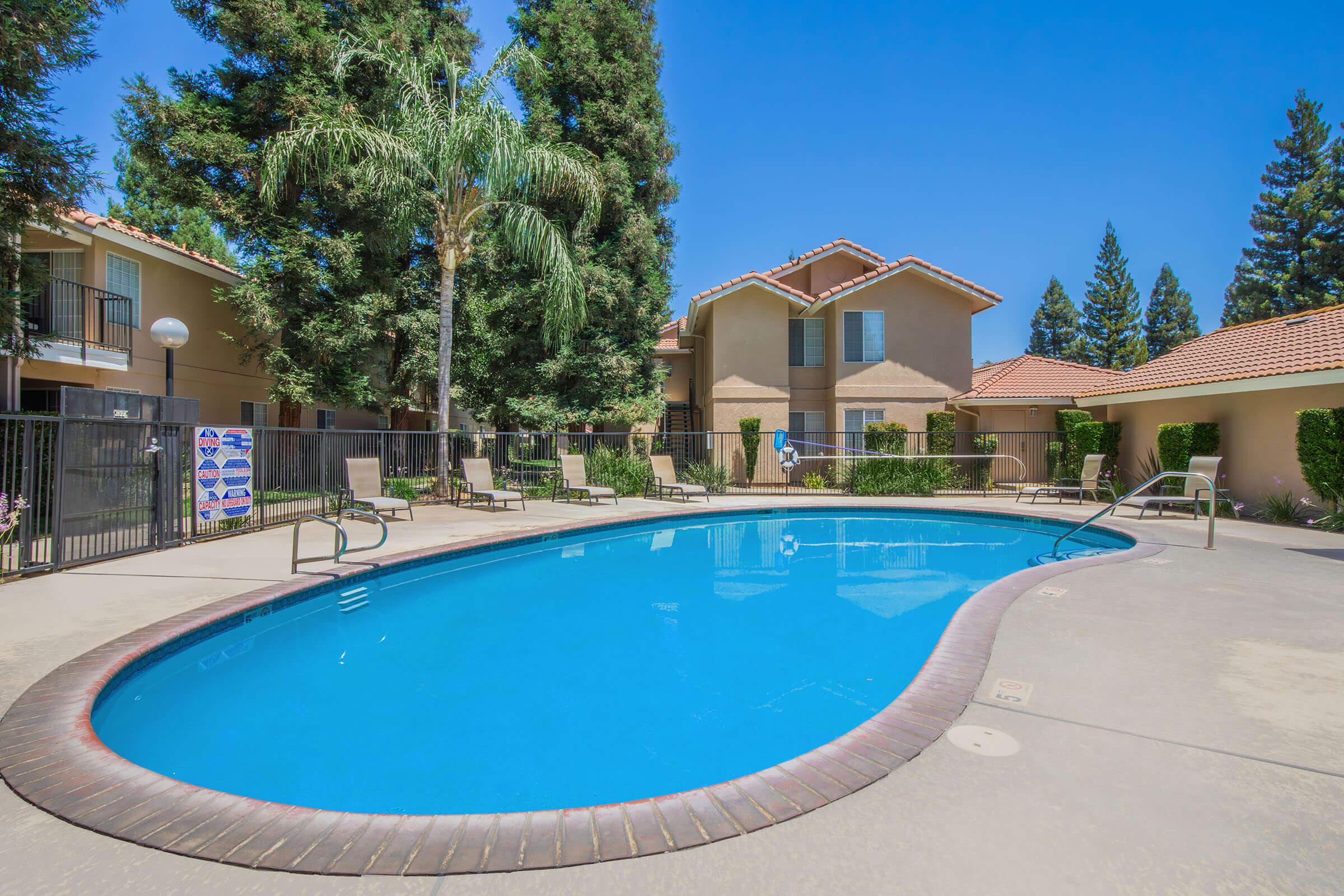 A clear blue swimming pool surrounded by lounge chairs and tall trees, with two multi-story apartment buildings in the background. The sky is bright and sunny, creating a vibrant outdoor scene. Fencing surrounds the pool area for safety.