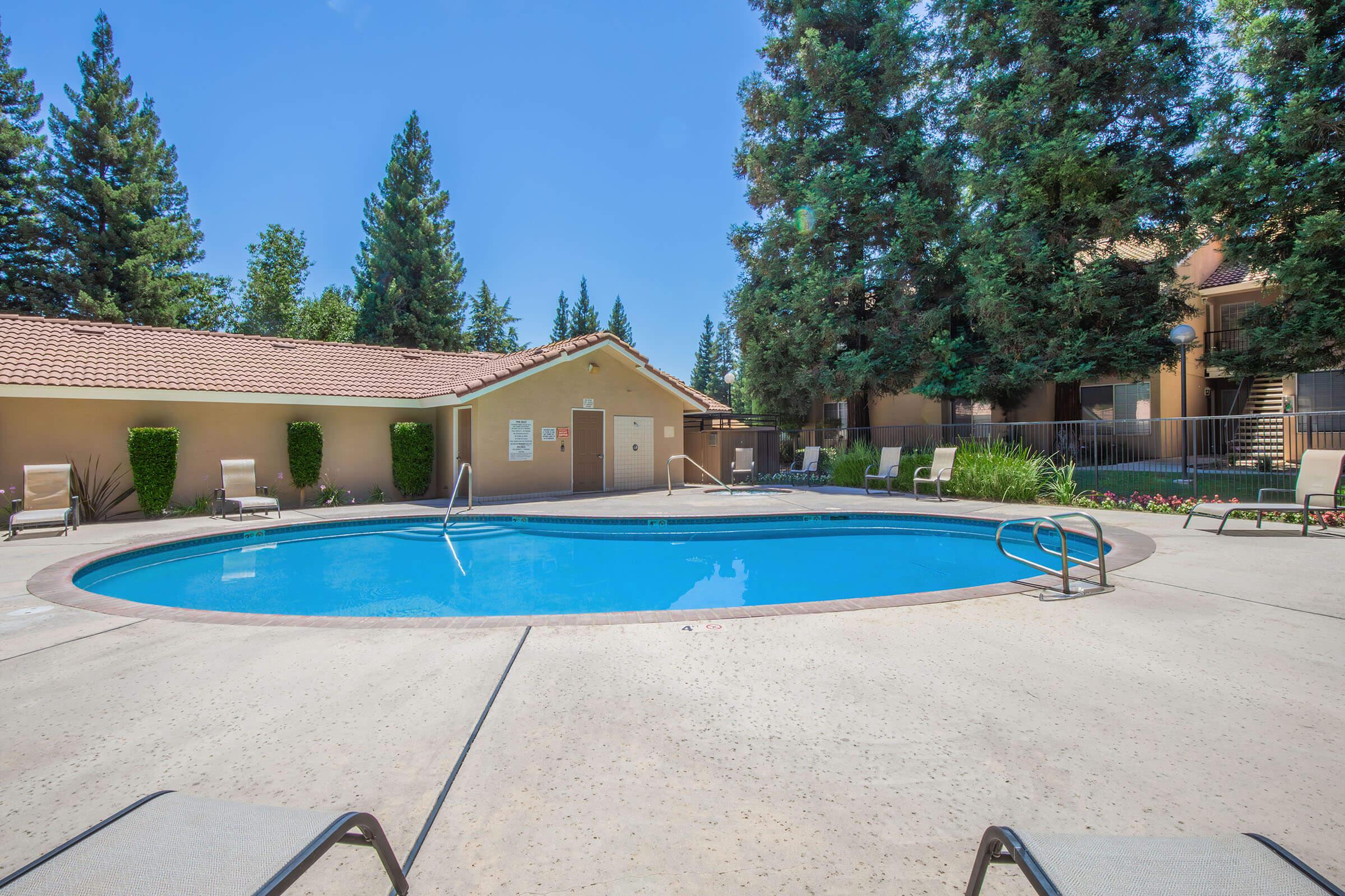 A sunny pool area featuring a round swimming pool surrounded by a concrete deck and lounge chairs. Lush green trees are in the background, and a small building with a roof is visible nearby. The scene is bright and inviting, ideal for relaxation and leisure activities.