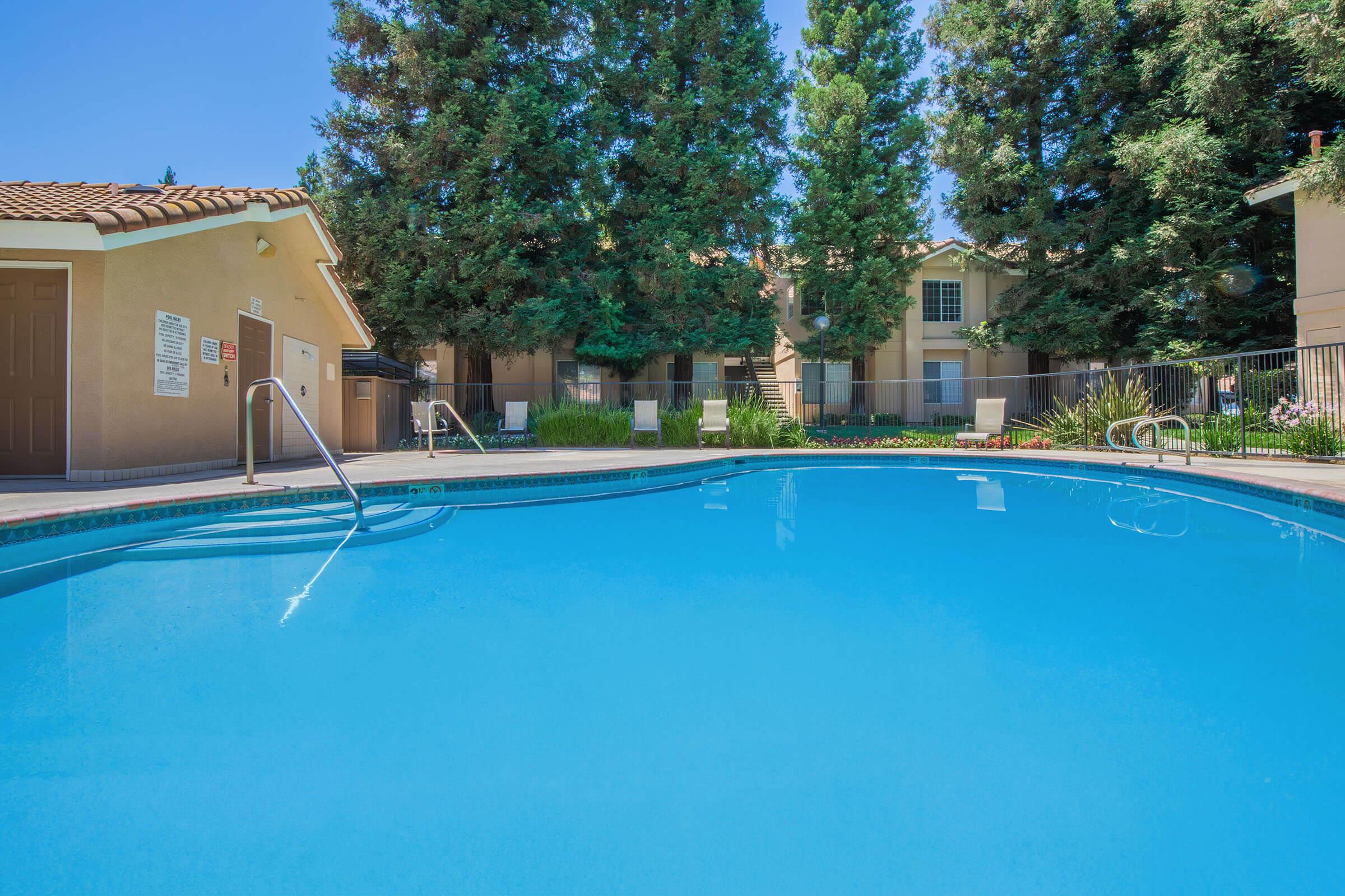A clear blue swimming pool surrounded by lush green trees and well-maintained landscaping. A few lounge chairs are positioned around the pool area. In the background, there are residential buildings with windows facing the pool, creating a relaxing atmosphere.