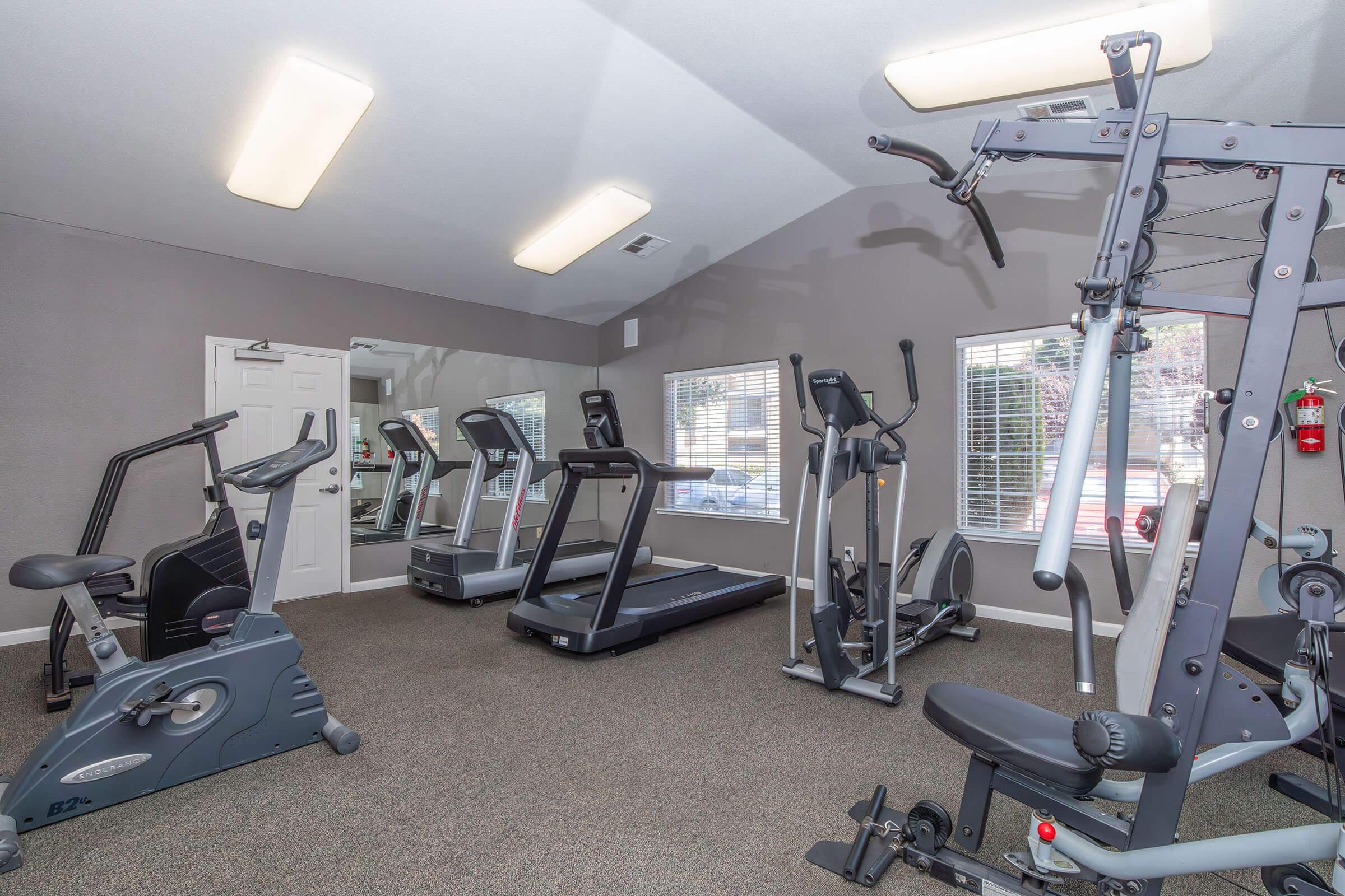 A spacious gym area featuring various exercise equipment. There are multiple treadmills and an elliptical machine, along with a multi-gym station. The room has large windows allowing natural light, with a neutral-colored wall and carpet flooring, creating a clean and inviting workout environment.