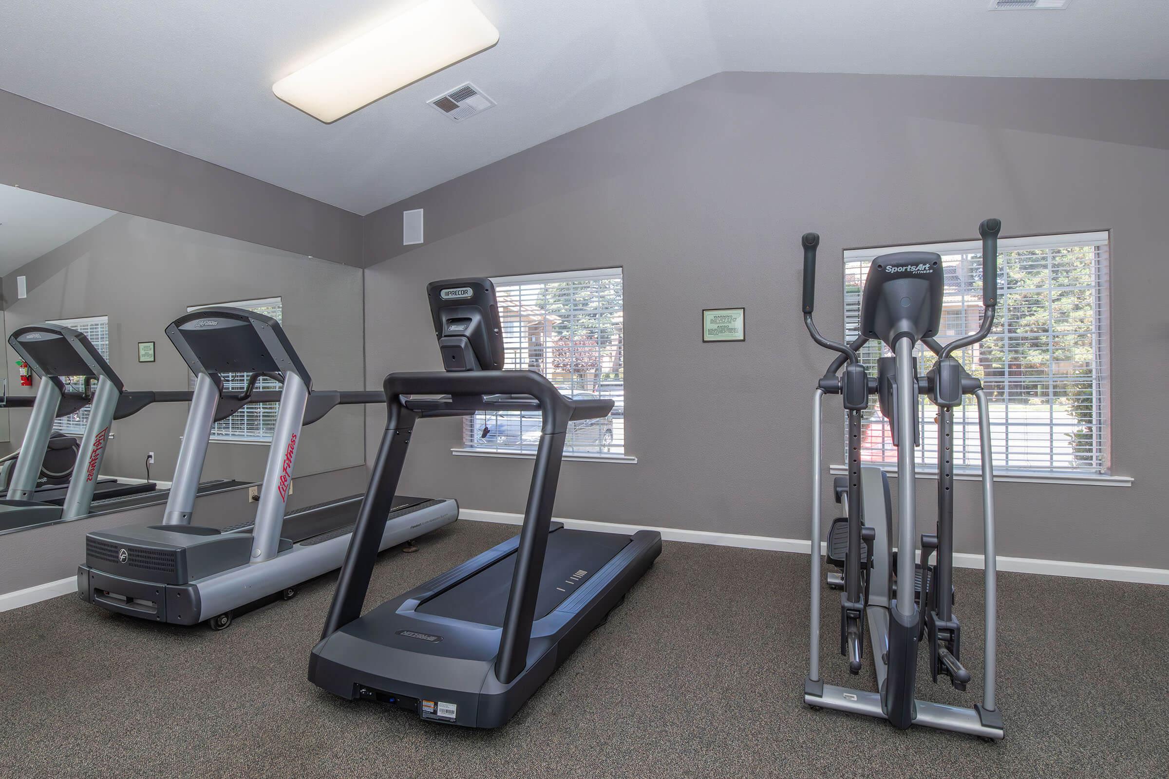 A fitness room featuring two treadmills and an elliptical machine. The space is well-lit with natural light coming through the windows and has a mirror on one wall. The flooring is carpeted, and the walls are painted in a neutral color.
