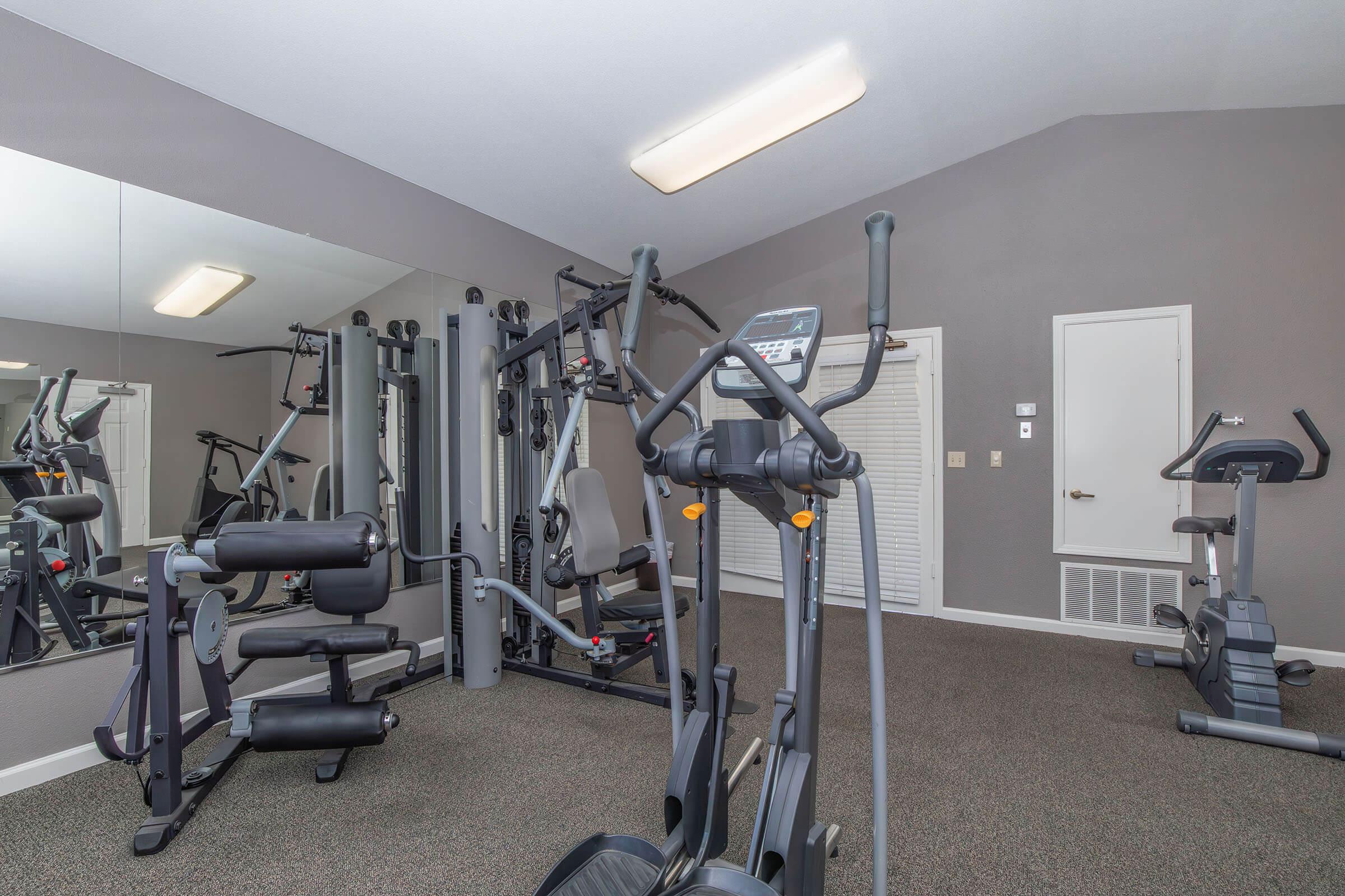 A modern gym interior featuring various exercise equipment, including a treadmill, elliptical cross-trainer, and weight machines. The space has gray walls, carpeted flooring, and mirrors that reflect the equipment. Natural light is limited, and the room appears well-organized and clean.