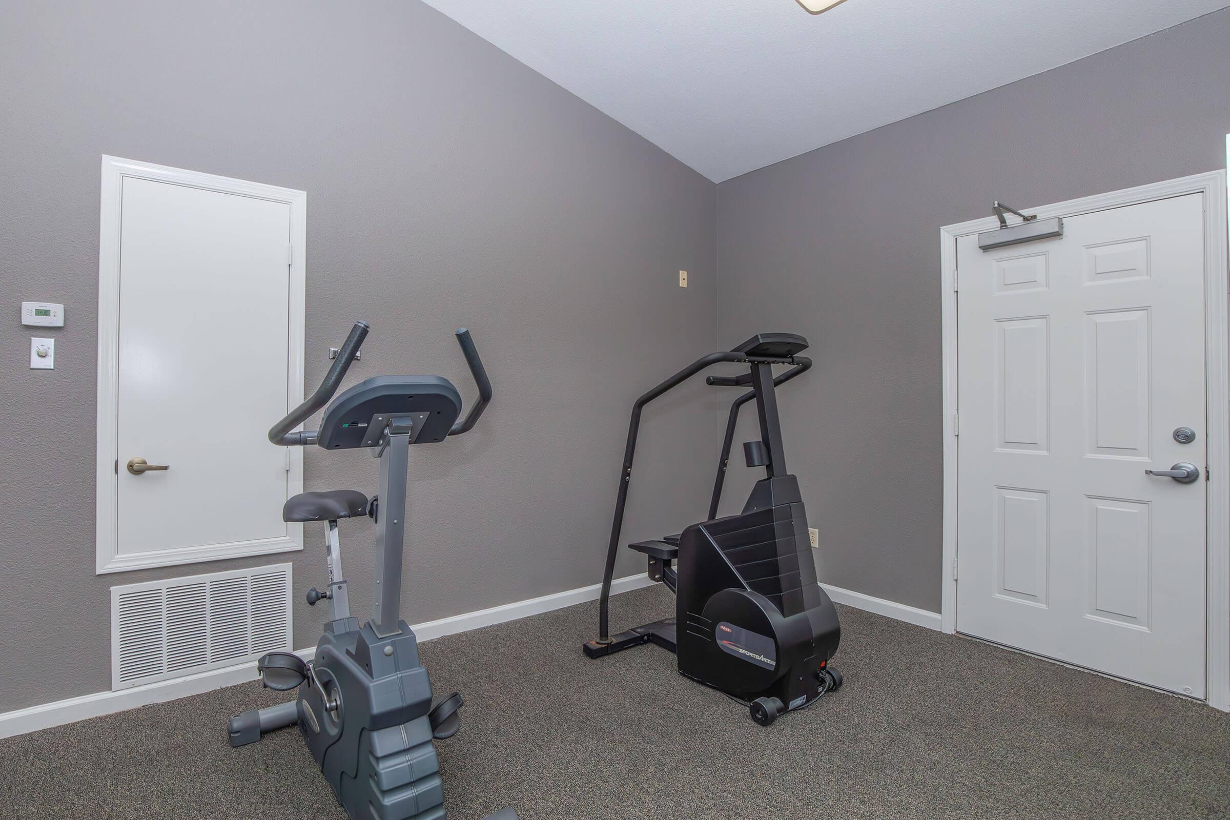 A small, empty home gym featuring a stationary exercise bike and an elliptical machine. The walls are painted grey, and there is a door leading outside. The floor is carpeted, and there is a heating vent on the wall.