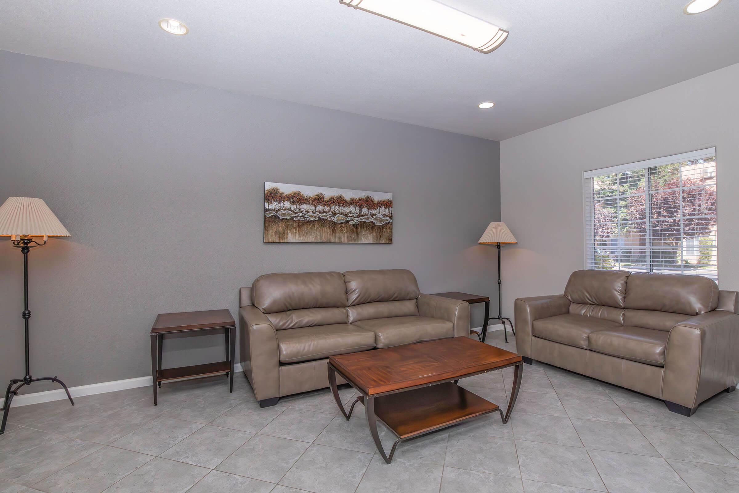 A modern living room featuring two brown leather couches, a wooden coffee table, and a small end table. There are two lamps providing lighting, and a landscape painting hangs on the wall. The room has gray walls and tile flooring, with natural light coming in from a window.