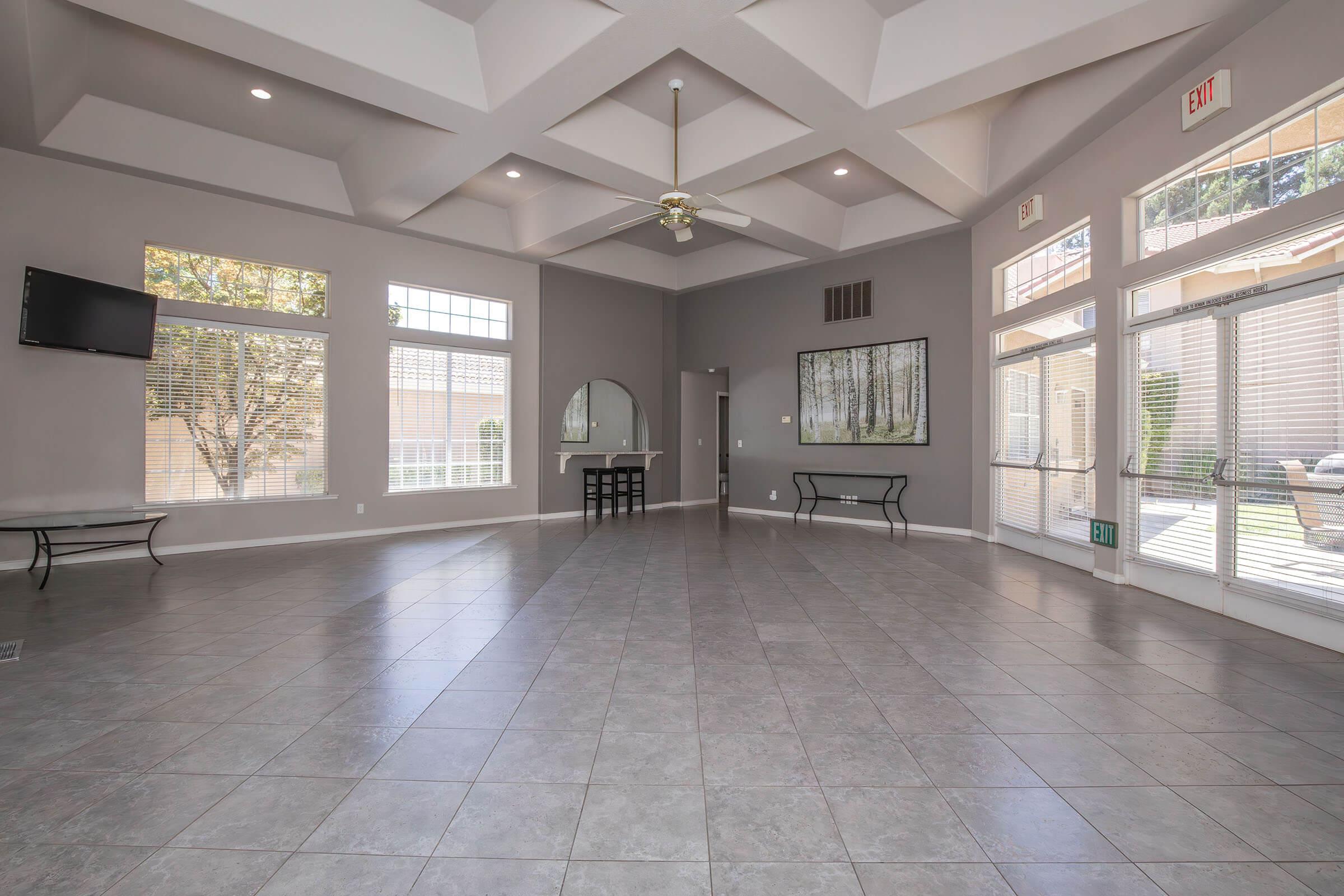 A spacious, well-lit interior room featuring a coffered ceiling, large windows allowing natural light, and tiled flooring. There is a flat-screen TV mounted on the wall, a round mirror above a console table, and a black bench against the wall, creating an inviting atmosphere.