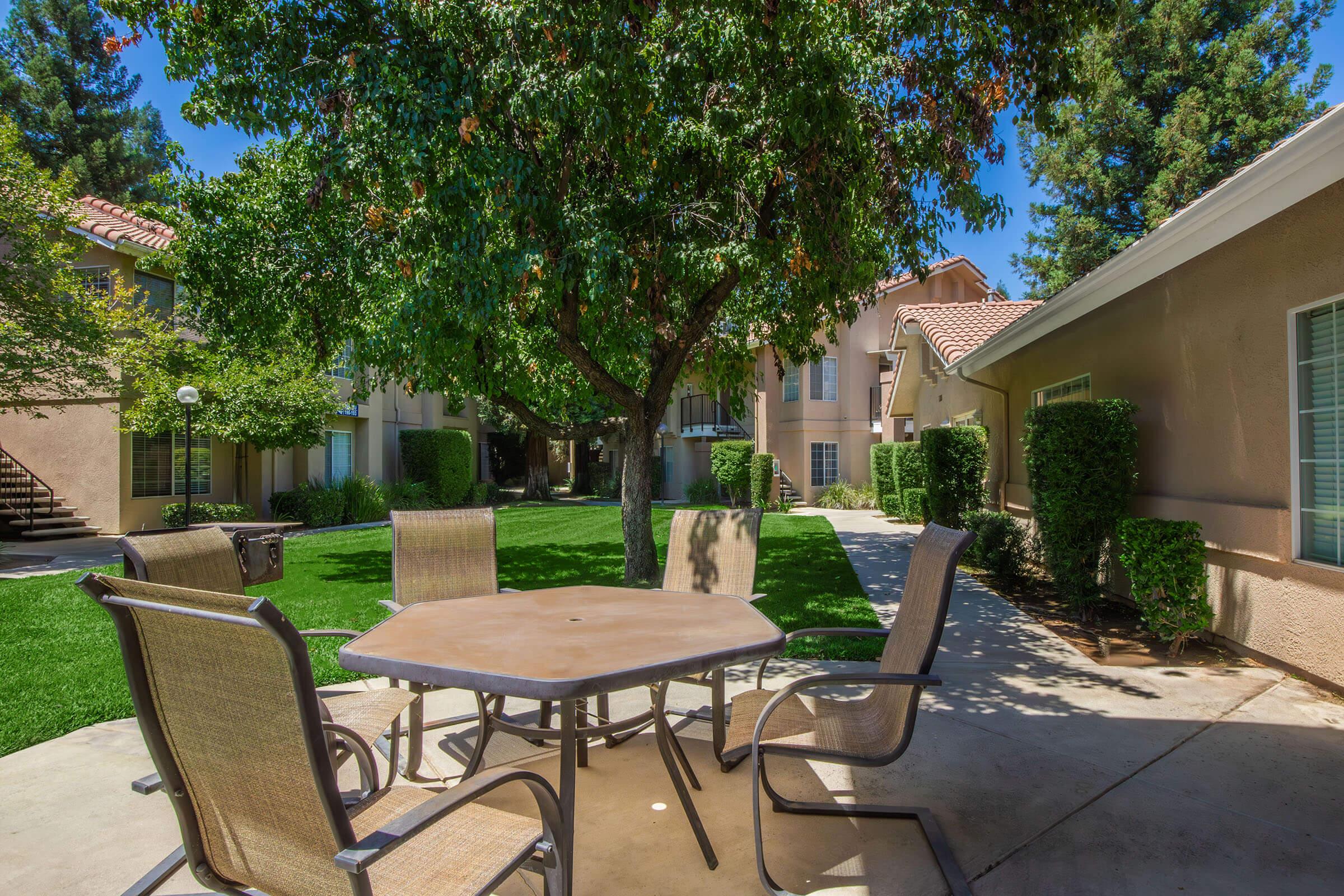 Outdoor patio area with a round table and four chairs under a large tree. The setting features well-maintained grass and buildings in the background, typical of a residential complex. Clear blue sky adds to the inviting atmosphere.