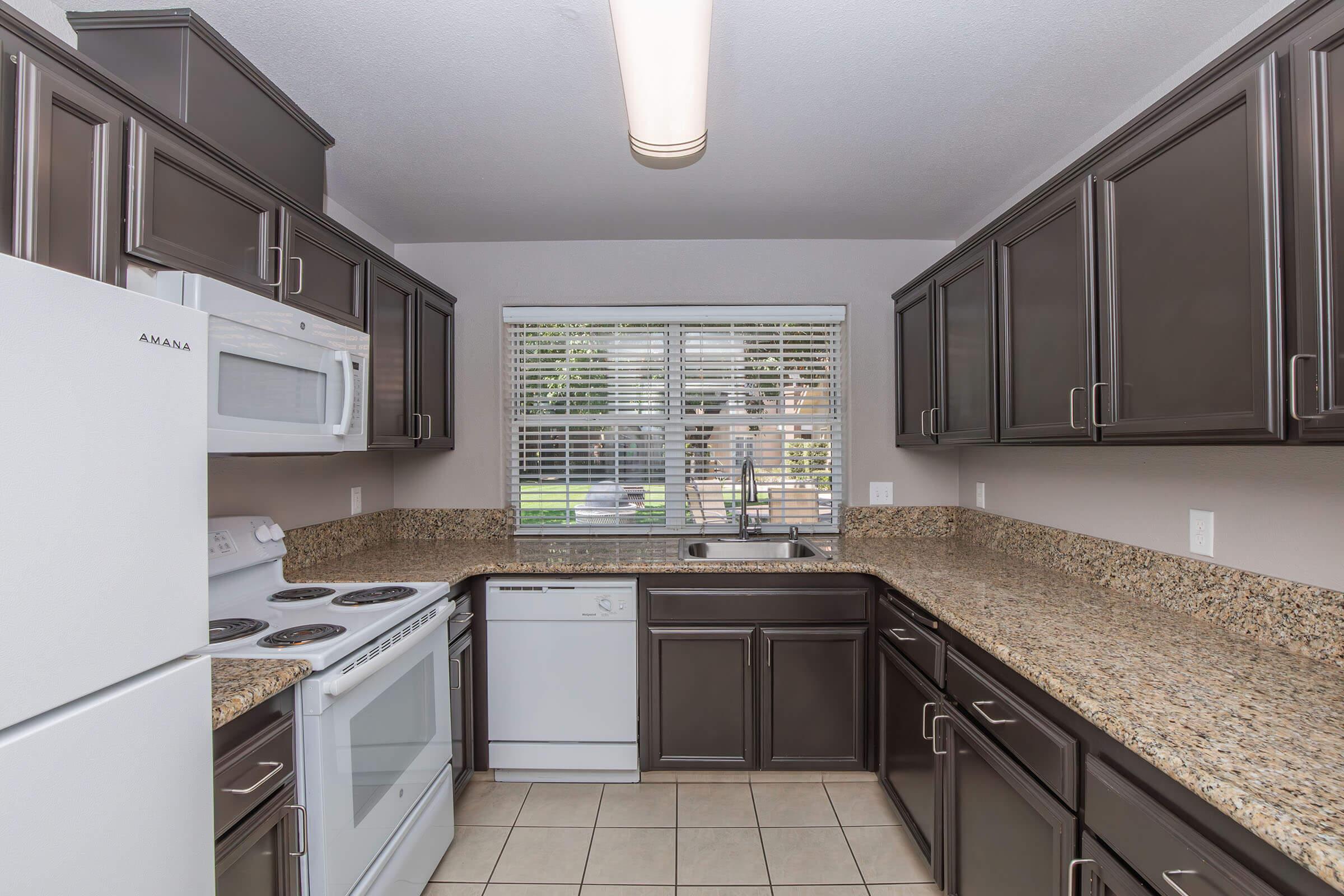 Modern kitchen featuring dark wood cabinets, granite countertops, and stainless steel appliances. Includes a white stove, dishwasher, and a window with natural light. The floor is tiled, and the overall design has a clean and organized look.