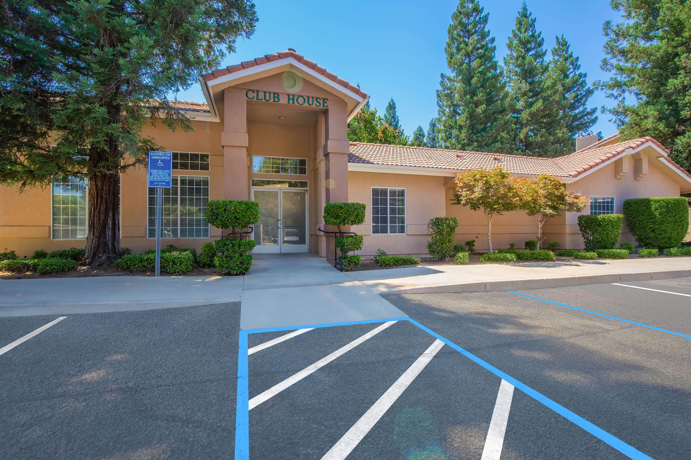 A modern clubhouse building with a sloping roof, large windows, and a welcoming entrance. Surrounded by neatly trimmed hedges and trees, the parking lot features designated spots, including accessible parking. The clear blue sky adds to the bright and inviting atmosphere.