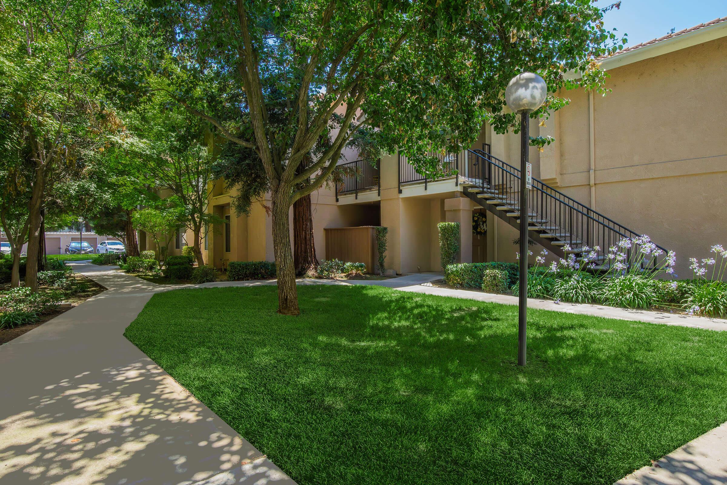A well-maintained courtyard featuring lush green grass, trees, and colorful shrubs. A paved walkway leads through the area toward a staircase that ascends to an upper level of a building. Soft sunlight filters through the foliage, creating a serene, inviting atmosphere.