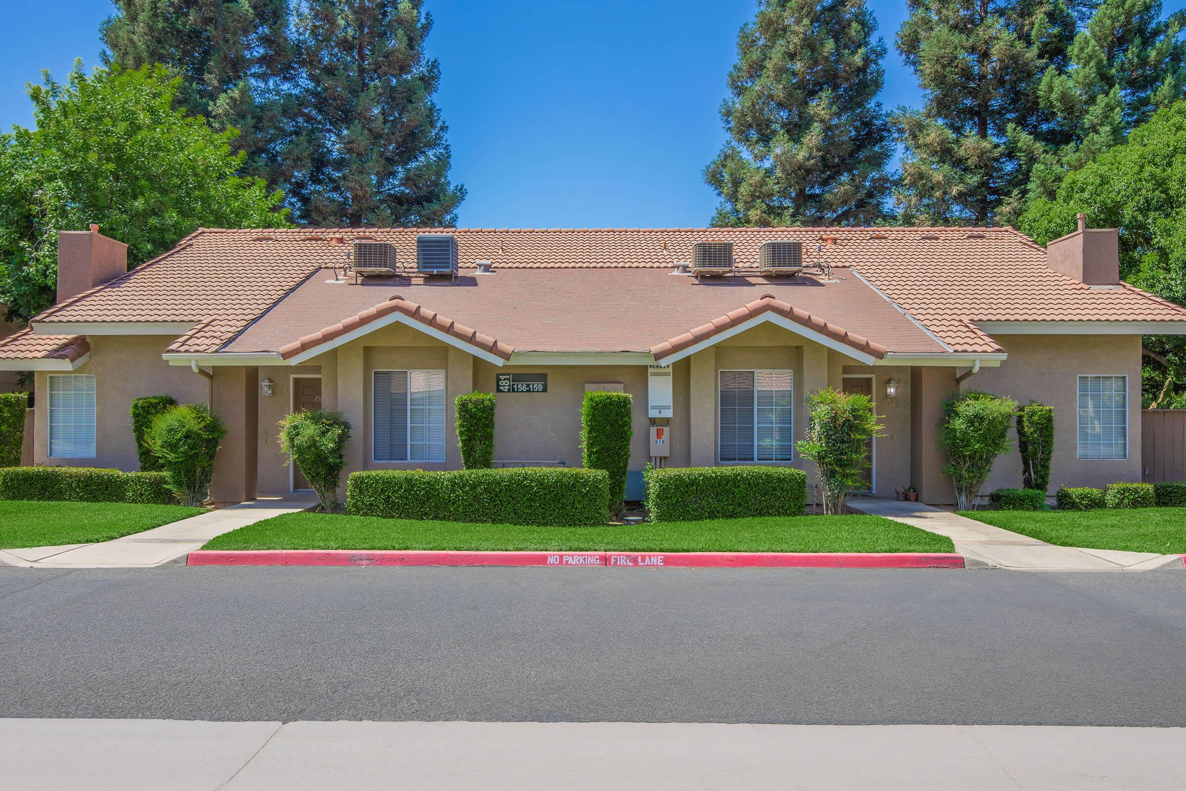 A duplex with a tan exterior and a red-tiled roof, featuring two front doors flanked by neatly trimmed bushes. The building is surrounded by green trees and a clear blue sky, with a well-maintained lawn visible in front. A concrete street runs in front of the property.