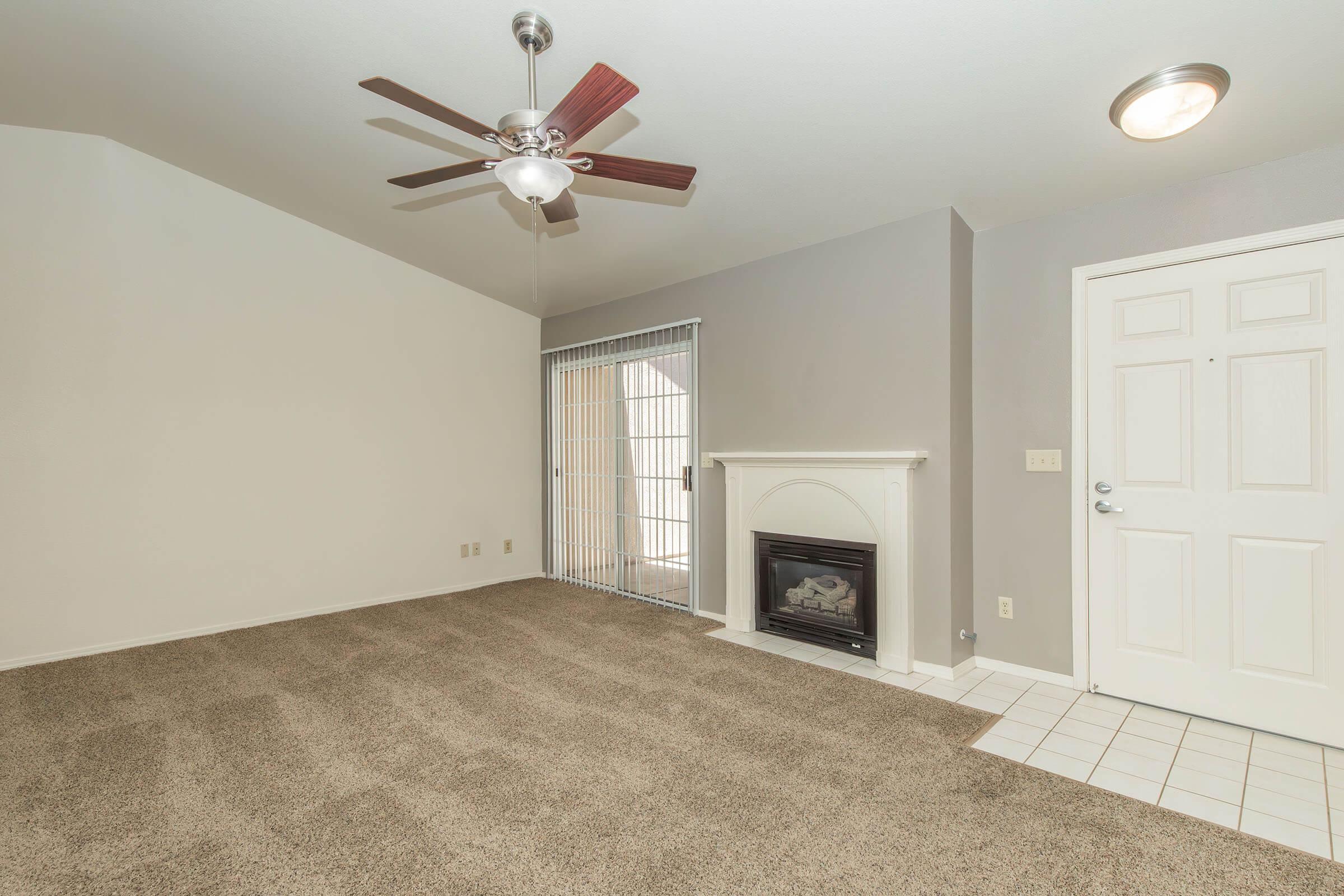 Spacious living room featuring a ceiling fan, light-colored walls, and beige carpet. A white fireplace is located against one wall, while large windows with vertical blinds let in natural light. The entrance door is visible on the right, with tile flooring near the doorway.