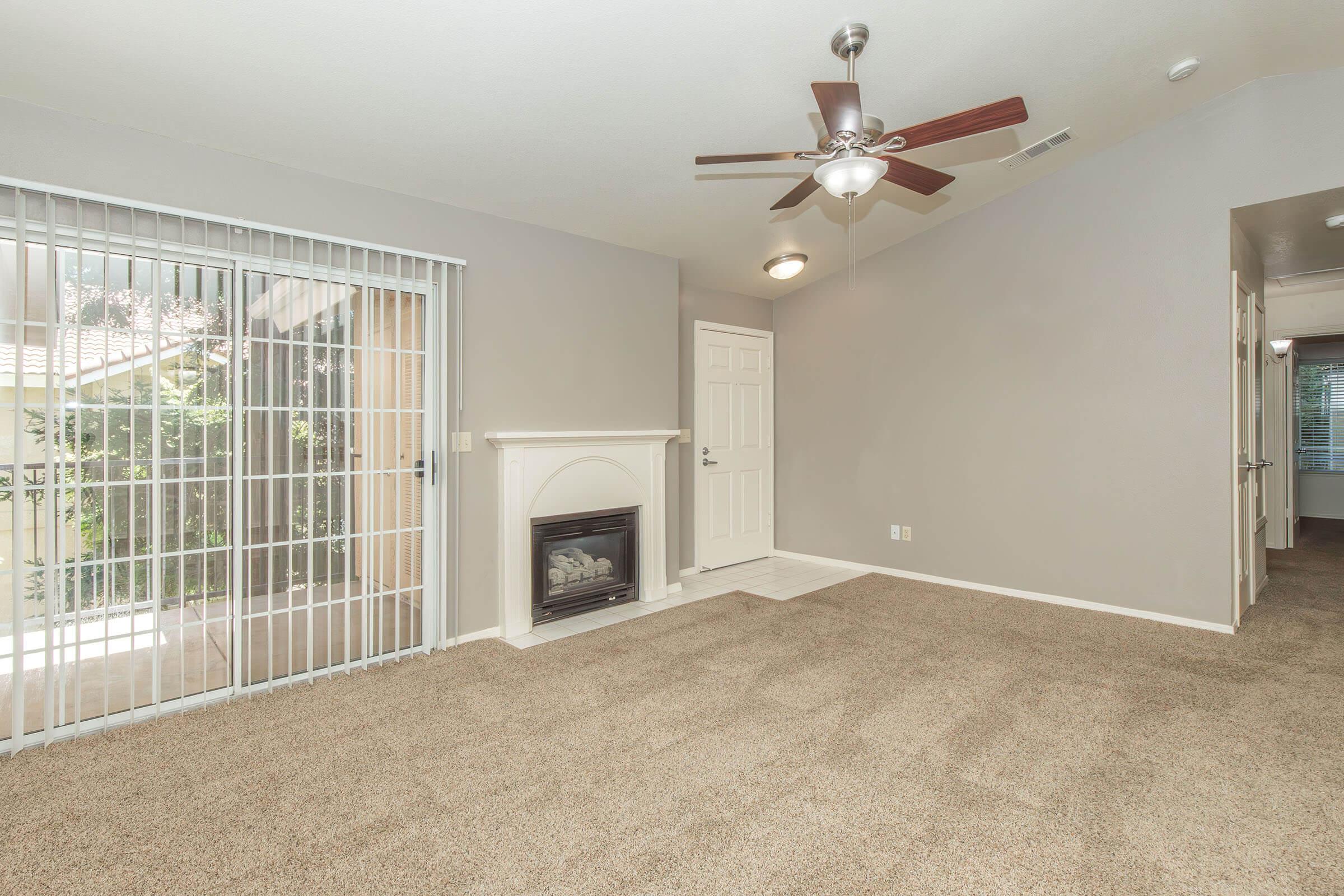 A spacious living area featuring a light gray wall, beige carpet, a ceiling fan, and a decorative fireplace. Large sliding glass doors provide natural light, leading to an outdoor space. The room has an open layout with a doorway visible on the right, suggesting access to other areas of the home.