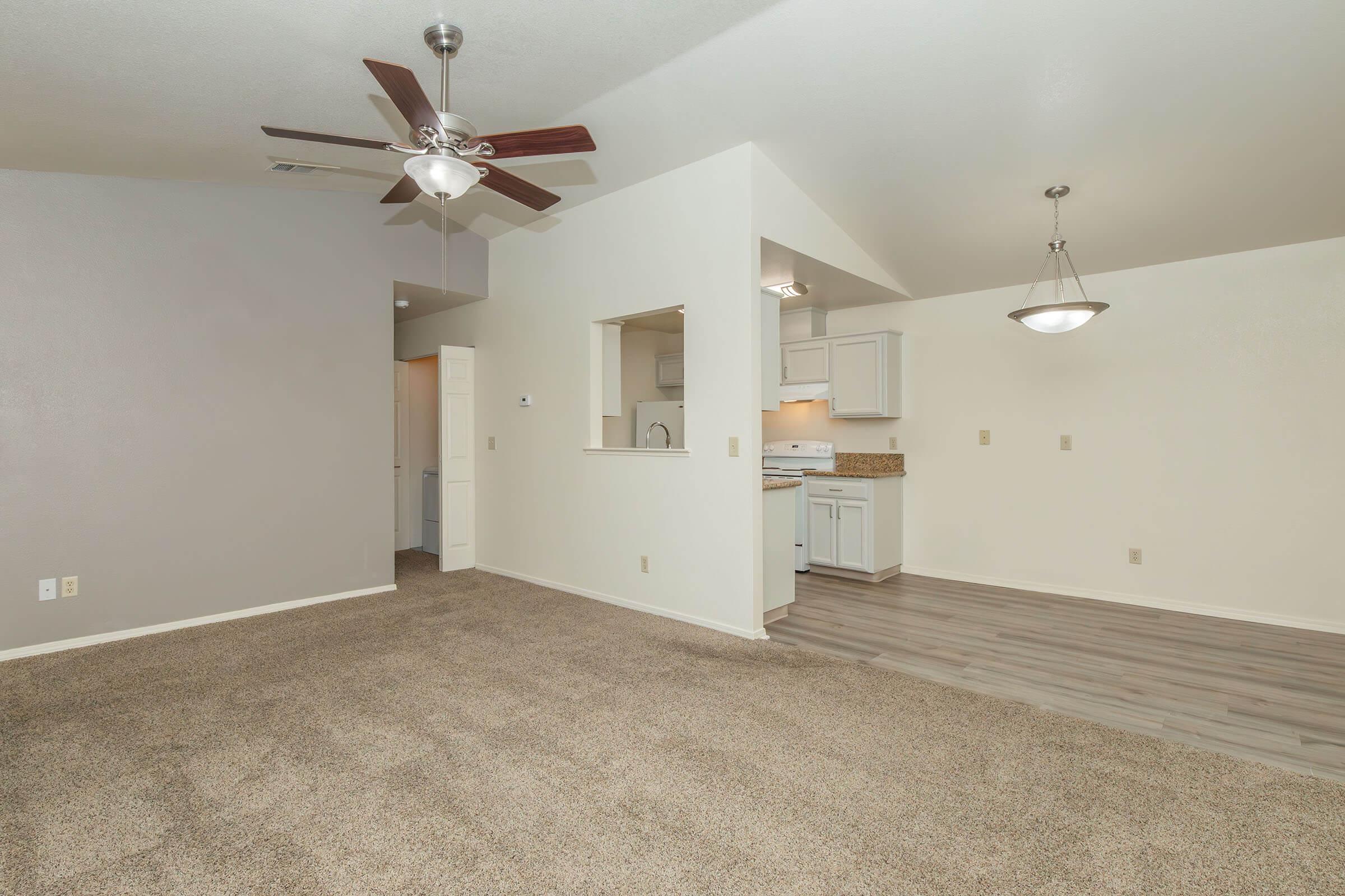 A spacious and brightly lit living area featuring a ceiling fan, neutral-colored walls, light brown carpet, and an open connection to the kitchen. The kitchen area has white cabinets and a countertop, creating a welcoming atmosphere.