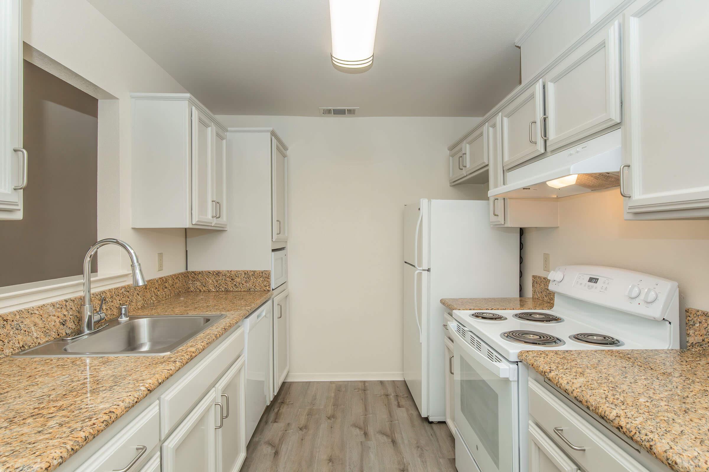 A modern kitchen featuring white cabinetry, granite countertops, an under-mount sink, and a white refrigerator. The kitchen also includes a white stove and oven with a range hood. The flooring is light-colored, and the space is well-lit with overhead lighting.