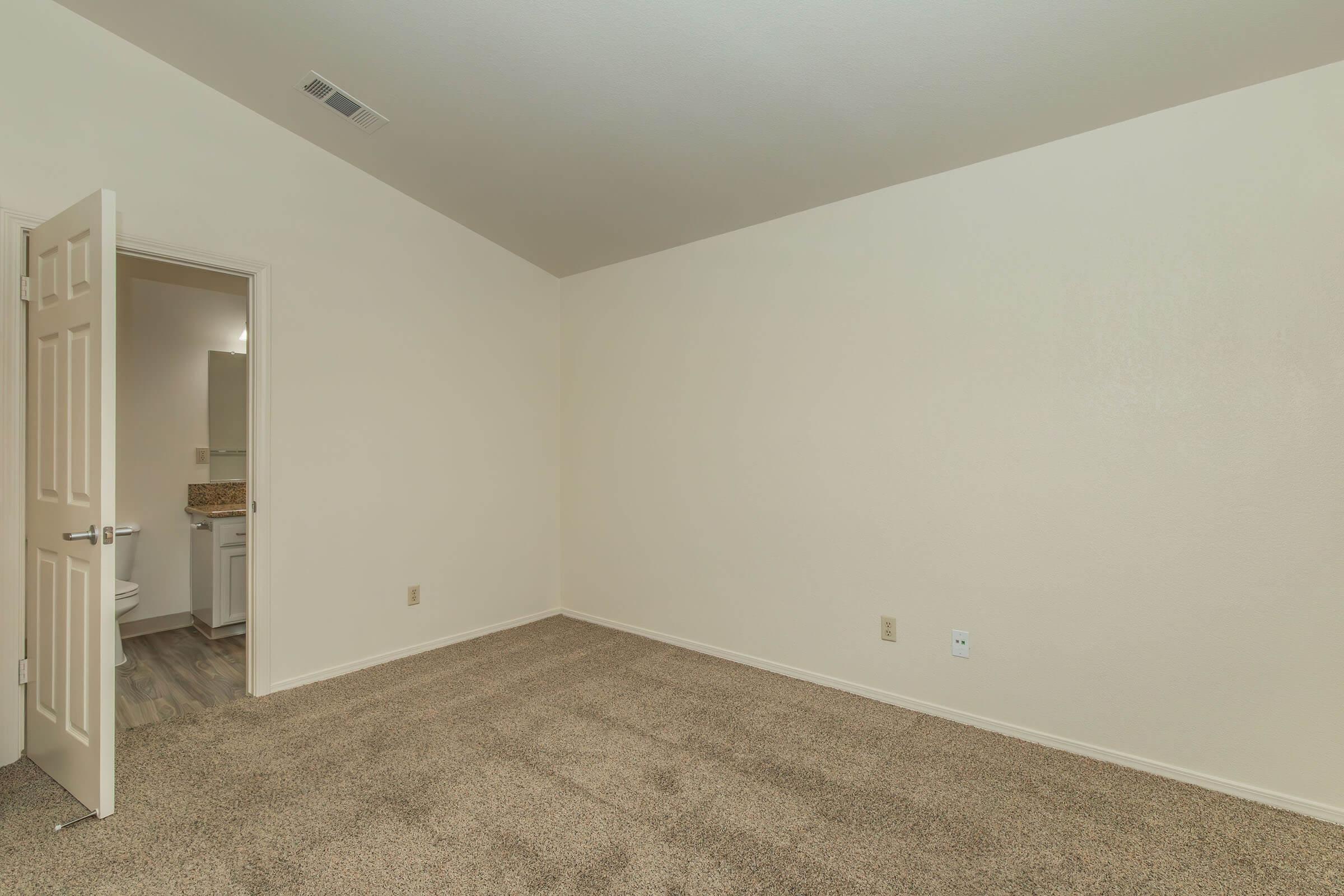A spacious, empty room with beige walls and light brown carpet. One wall features an open door leading to a bathroom area. The room has no furniture, creating a minimalist atmosphere. The ceiling has a slight slope, and there is a small area of light reflecting off the walls.
